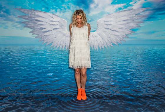 Een vrouw in een witte jurk en oranje laarzen staat op een geschilderd wateroppervlak met grote witte engelen_vleugels achter haar.