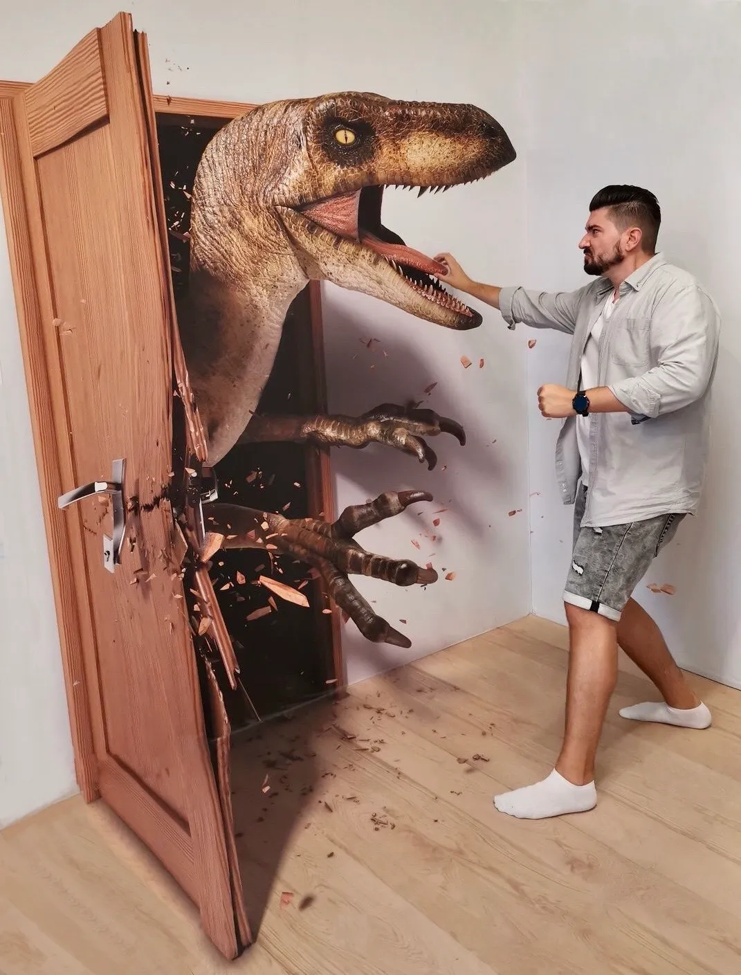 Een man in een licht hemd en korte broek reikt naar een 3D-dinosaurus die uit een houten deur komt, met rondvliegend puin.