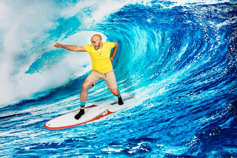 Een man in een geel shirt en beige korte broek die op een surfplank staat voor een grote 3D-golfallusie