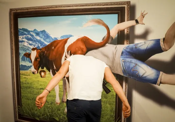 Persoon in wit shirt en donkere broek staand voor een groot 3D-illusieschilderij van een koe en bergen, met de benen van een andere persoon die boven het kunstwerk lijken te zweven.