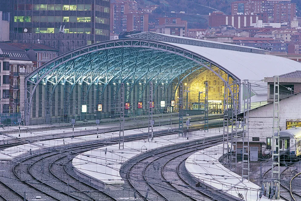 Widok z zewnątrz na stację kolejową Bilbao-Abando, ukazujący jej dużą, łukowatą konstrukcję szklanego dachu nad peronami oraz zakrzywione tory na pierwszym planie