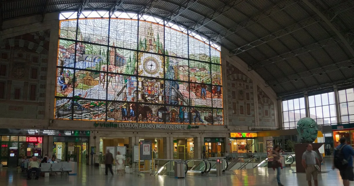 Wnętrze dworca kolejowego Bilbao-Abando z dużym łukowatym witrażem przedstawiającym sceny z miasta z centralnym zegarem, umieszczonym nad schodami ruchomymi i bramkami biletowymi, przez które przechodzą podróżni.
