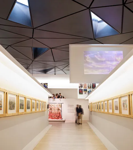 Long gallery hallway with geometric ceiling, framed artworks on white walls, and visitors viewing an exhibition