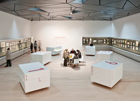 Large gallery space with white display cases, illuminated wall panels, and a geometric ceiling