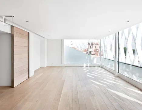 Minimalist interior space with light wood flooring, white walls, and large windows overlooking a cityscape.