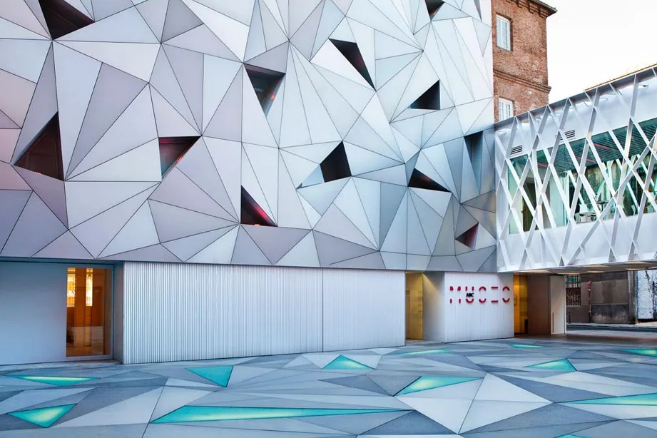 Modern building with a geometric white and gray facade, illuminated blue patterns on the ground, and a sign reading 'MUSEO' at the entrance.