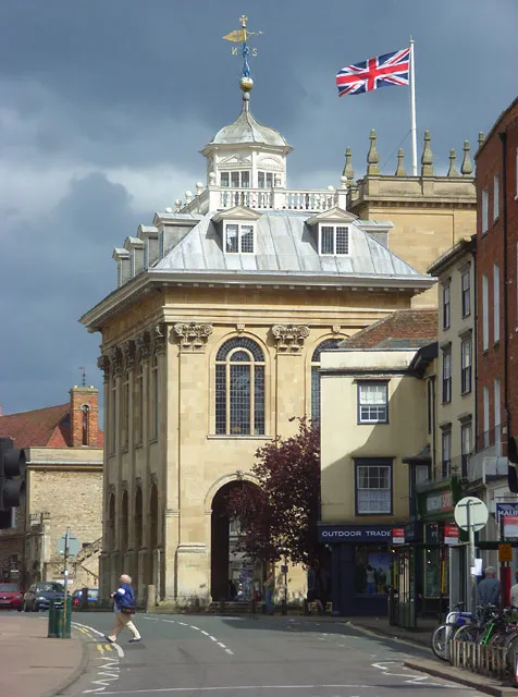 Vista exterior del Abingdon County Hall Museum que destaca por su fachada de piedra, un gran ventanal arqueado, una cúpula con veleta y una bandera del Reino Unido ondeando en el techo.