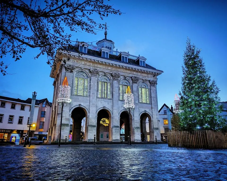 Edificio de piedra del ayuntamiento al atardecer con entrada arqueada en la planta baja, grandes ventanales, cúpula en el tejado, un árbol de Navidad a la derecha y una plaza empedrada en primer plano