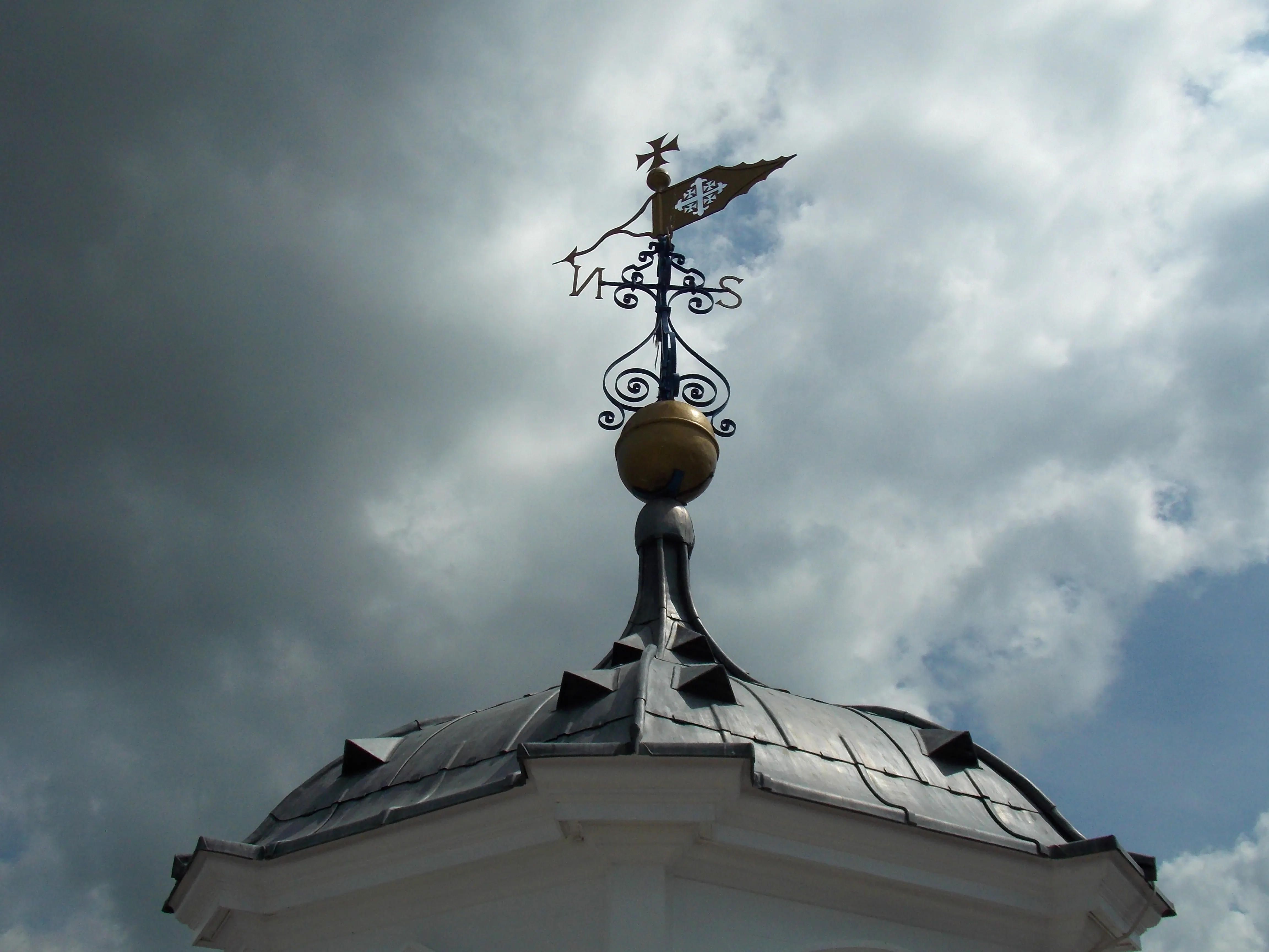 Veleta de hierro ornamentada con una bola dorada sobre una cúpula cubierta de plomo contra un dramático cielo nublado