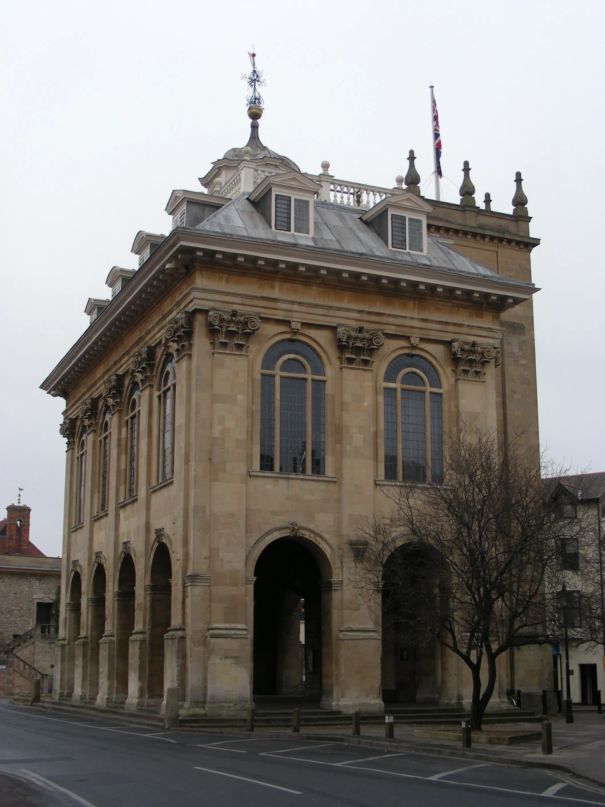 Stenen gevel van het Abingdon County Hall Museum met gewelfde openingen op de begane grond, hoge gewelfde ramen, decoratief metselwerk en een koepel met een windwijzer en een Union Jack-vlag.