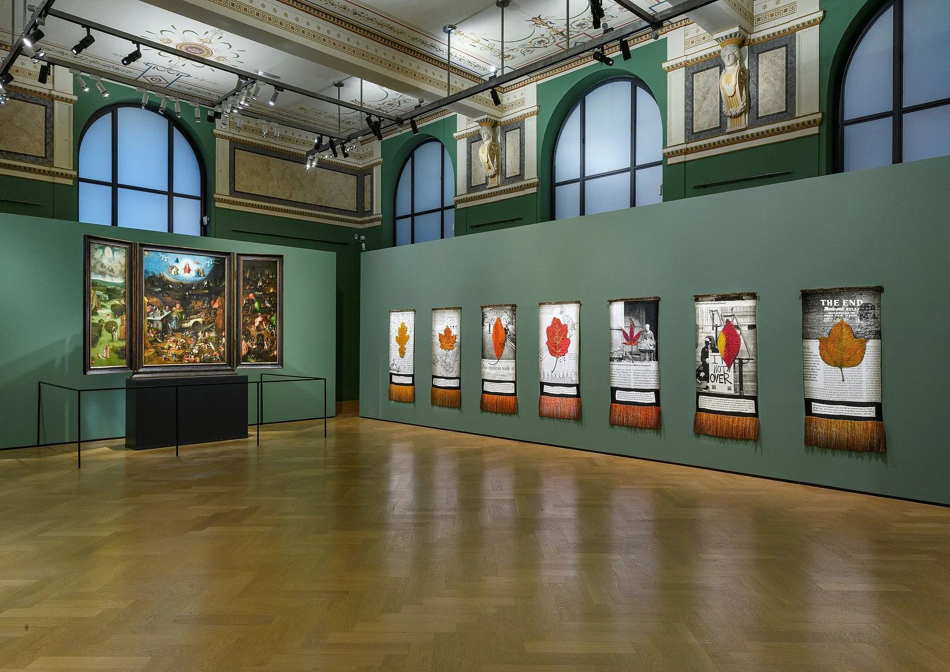 Gallery room with a large painting on the left and several framed artworks on a green wall under arched windows