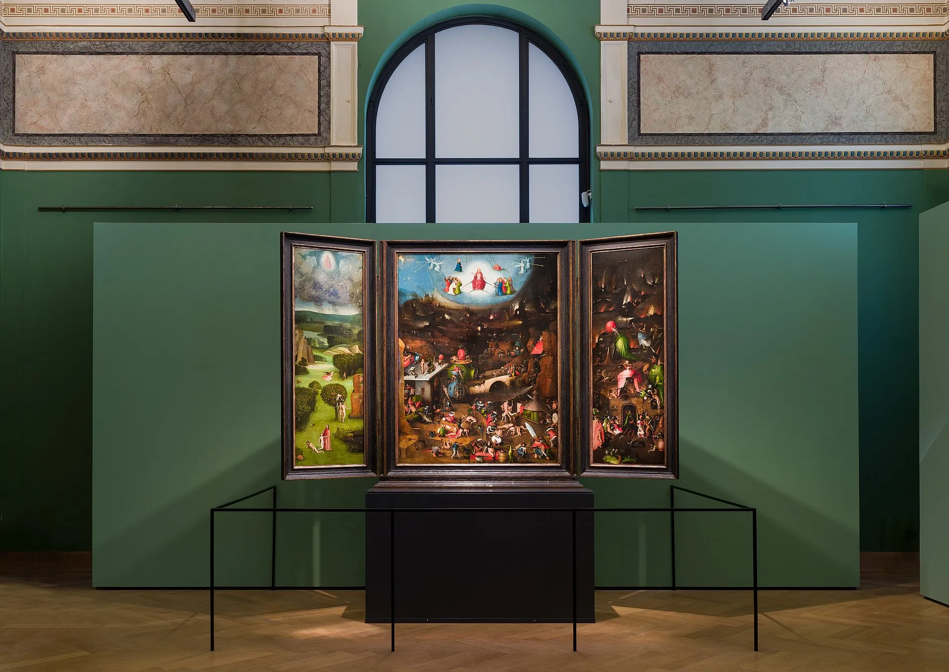 A triptych painting with three panels displayed on a black pedestal in a gallery with green walls and an arched window.