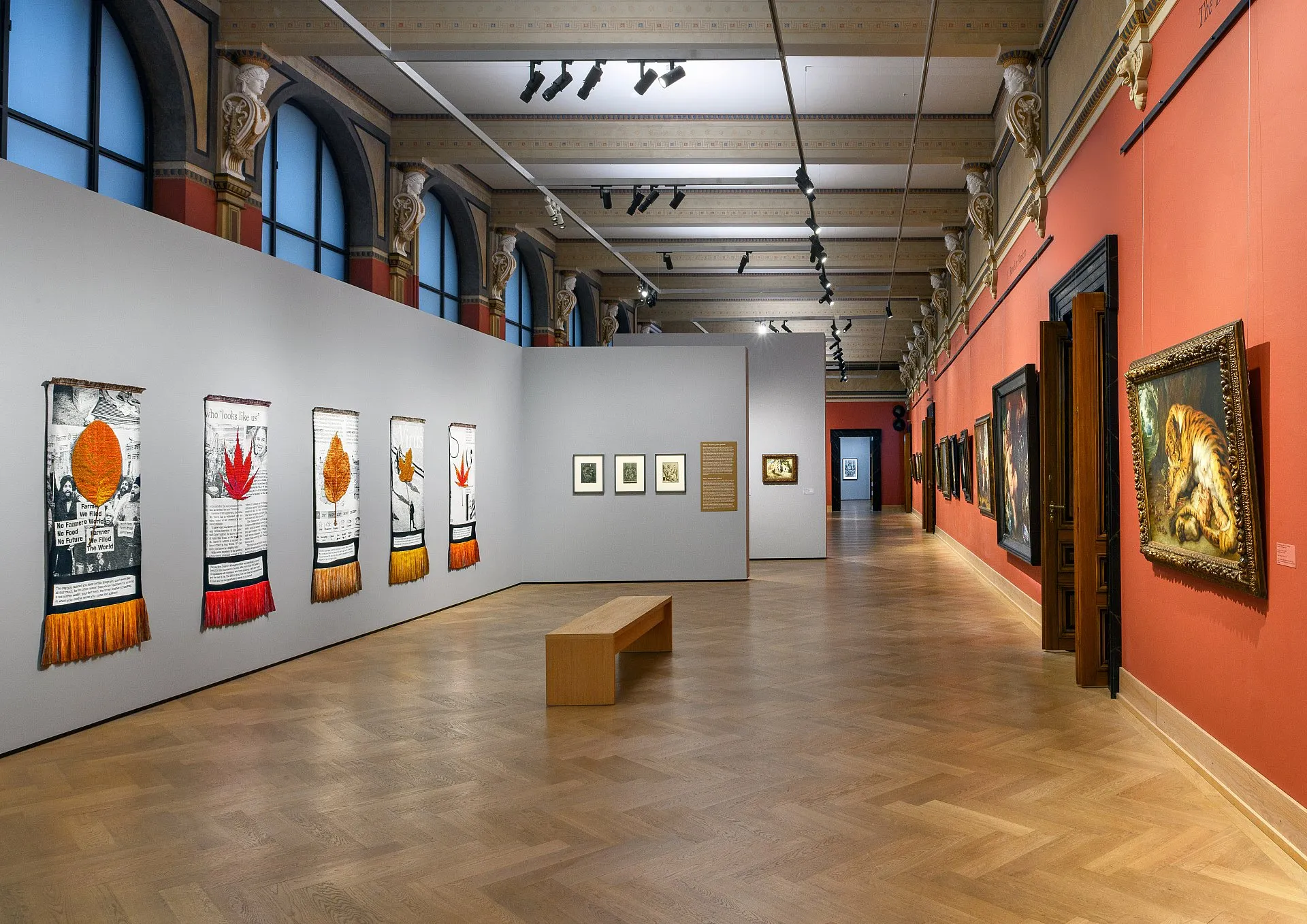 Long gallery hall with artworks on white and red walls, wooden floors, and arched windows