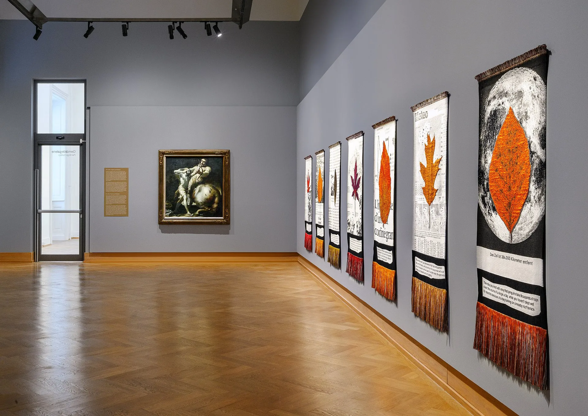 Art gallery with wooden floors and gray walls, displaying a framed painting and a row of textile artworks with orange leaf patterns