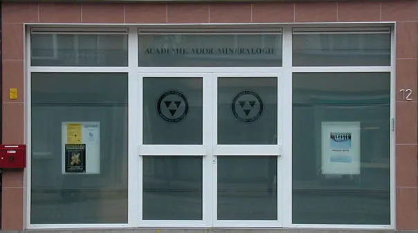 Entrada acristalada del ACAM Mineralogical Museum con puertas dobles de marco blanco, dos emblemas circulares en los paneles superiores, carteles en las ventanas laterales y un buzón rojo a la izquierda