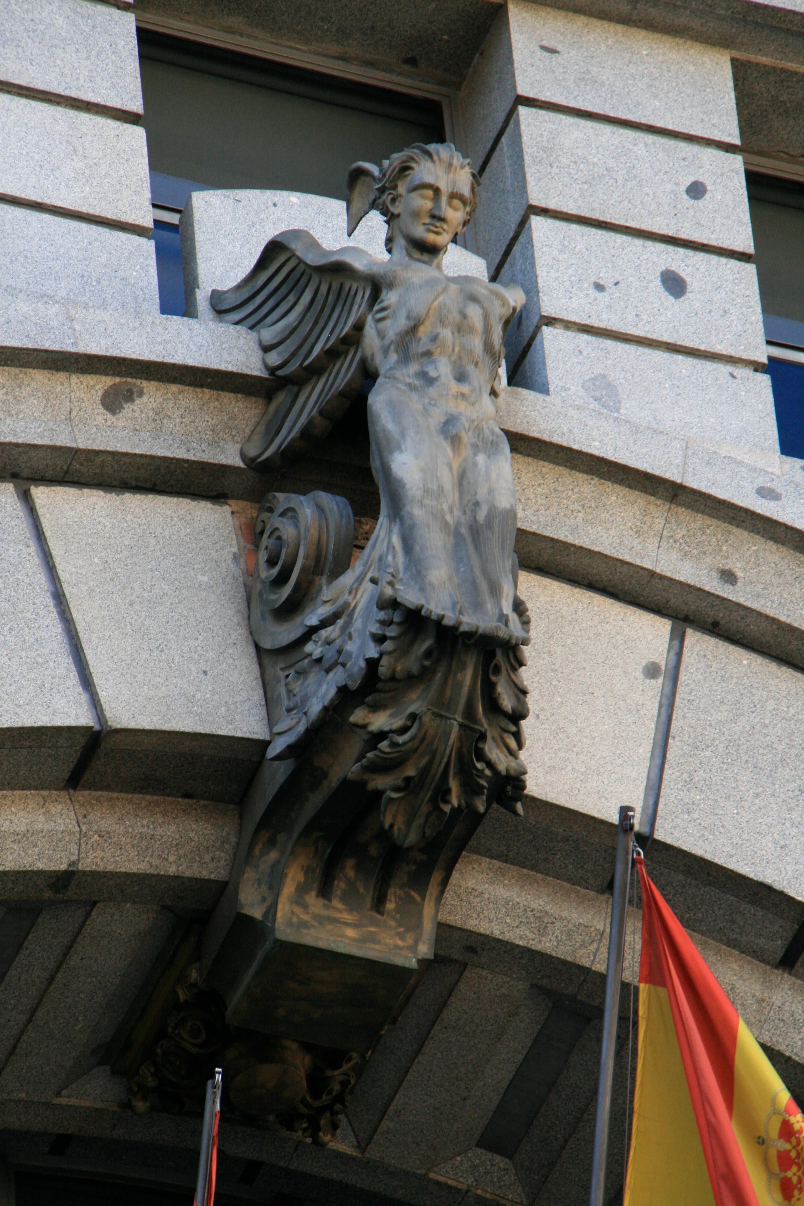 Figura alada esculpida en la fachada de un edificio de piedra con una bandera española