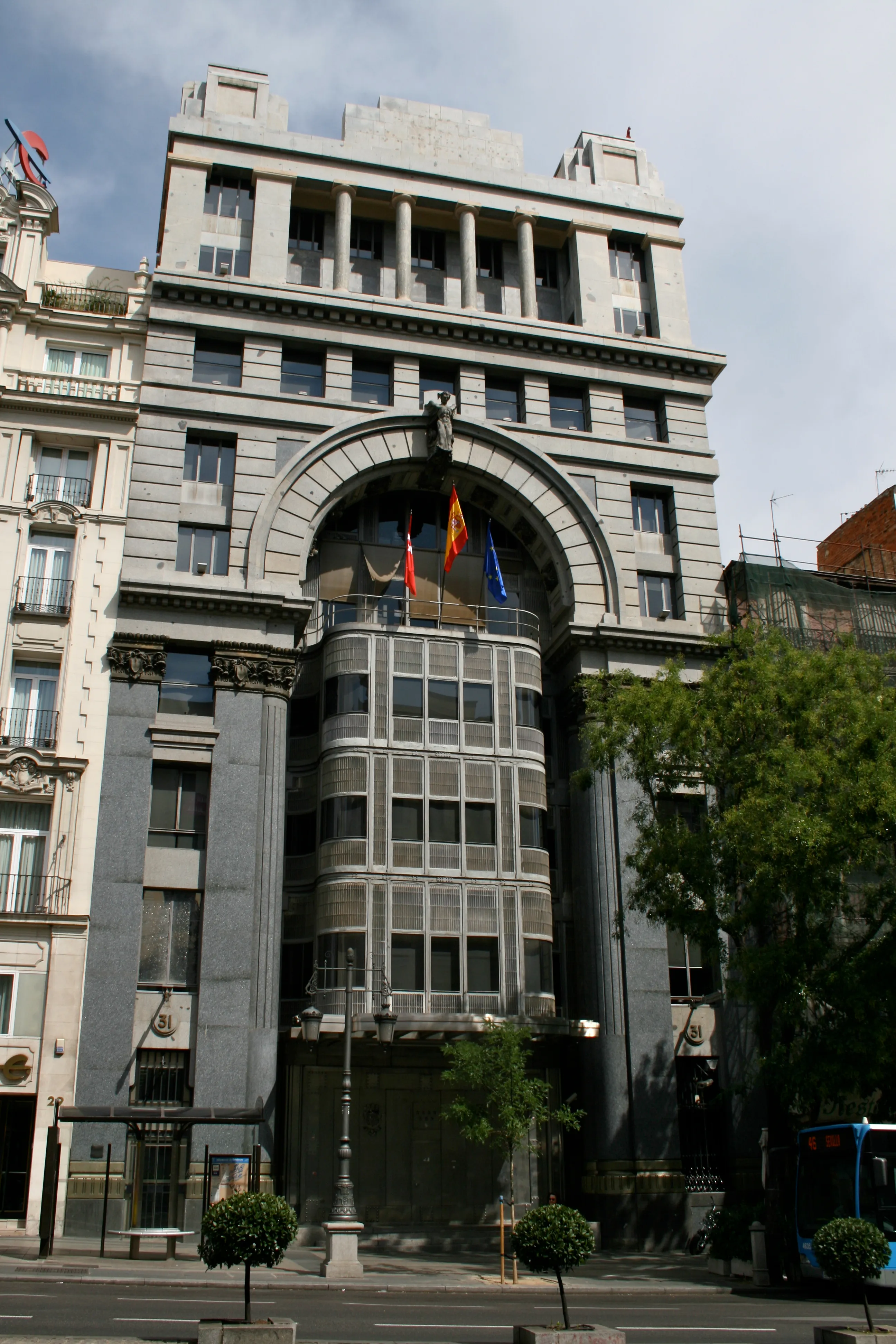 Un edificio majestuoso con una gran entrada arqueada, columnas clásicas y una moderna estructura de ascensor de vidrio, con banderas colgando sobre la entrada.
