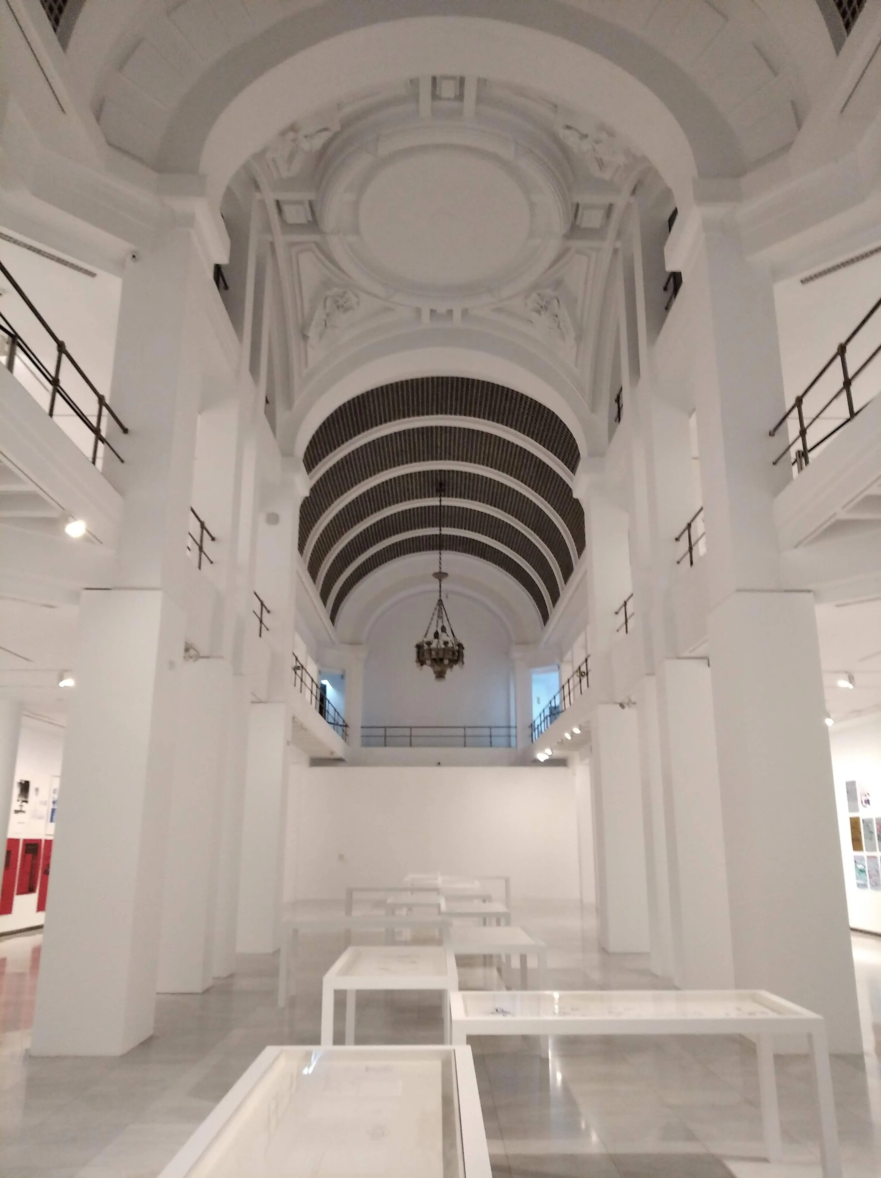Techo alto abovedado con detalles ornamentados y una lámpara de araña en un espacio de galería blanco con mesas de exhibición