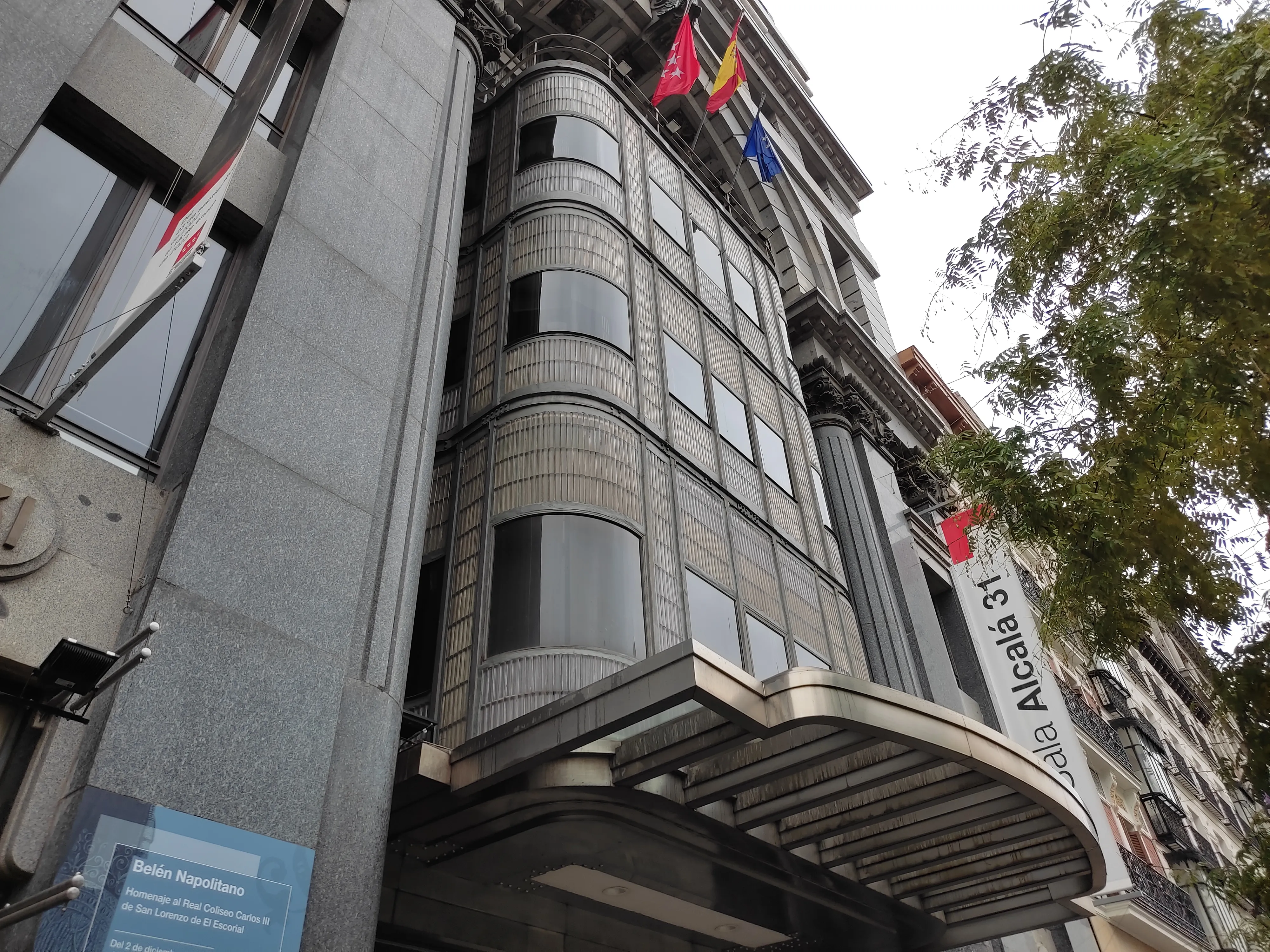 El exterior de Sala Alcalá 31, un edificio con fachada de vidrio curvo y un letrero que dice 'Sala Alcalá 31' sobre la entrada, con banderas ondeando en el tejado.