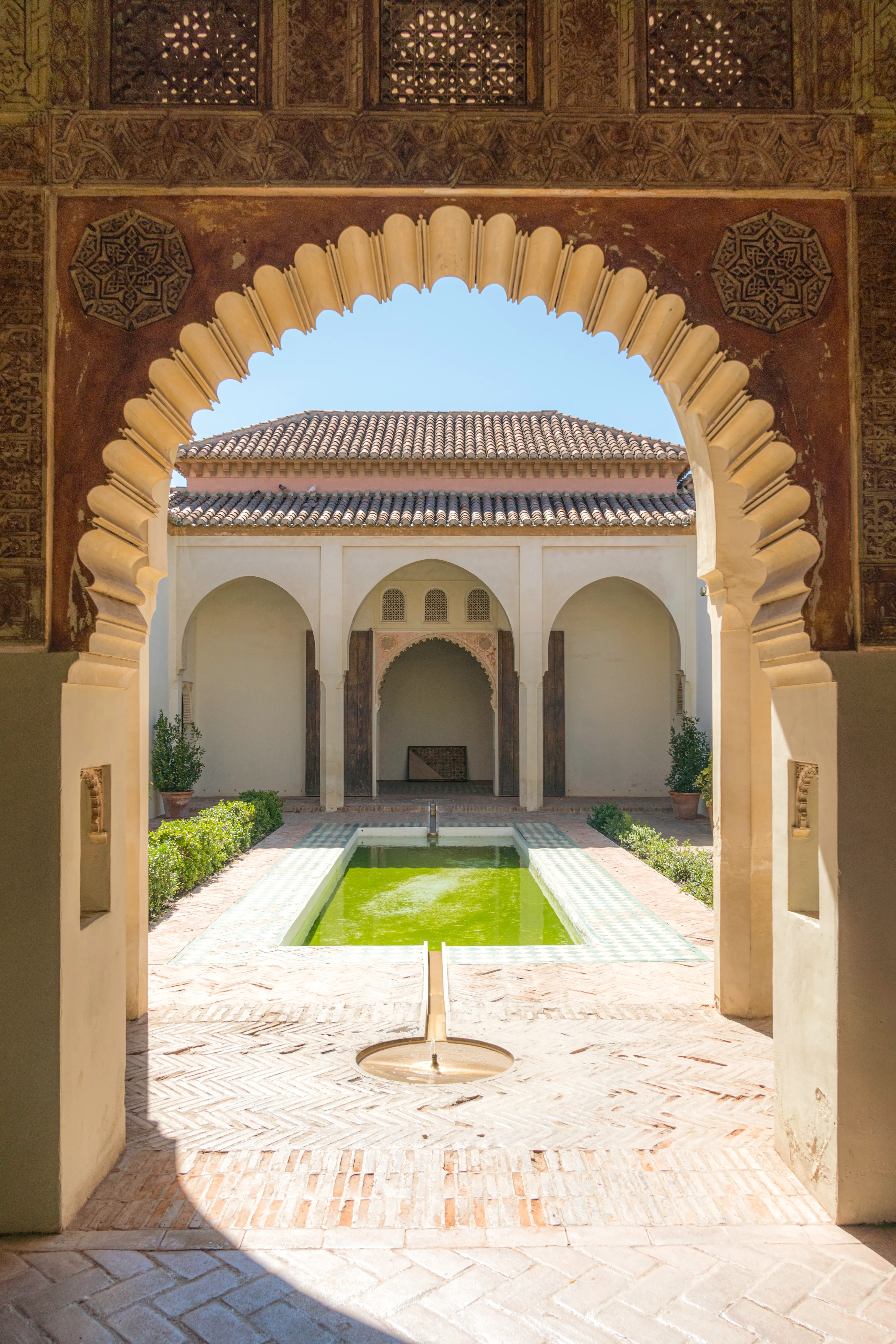 Arco de herradura de estilo nazarí que enmarca un estanque rectangular en el Patio de la Alberca de la Alcazaba de Málaga, con muros encalados y setos verdes bajo un cielo azul despejado.