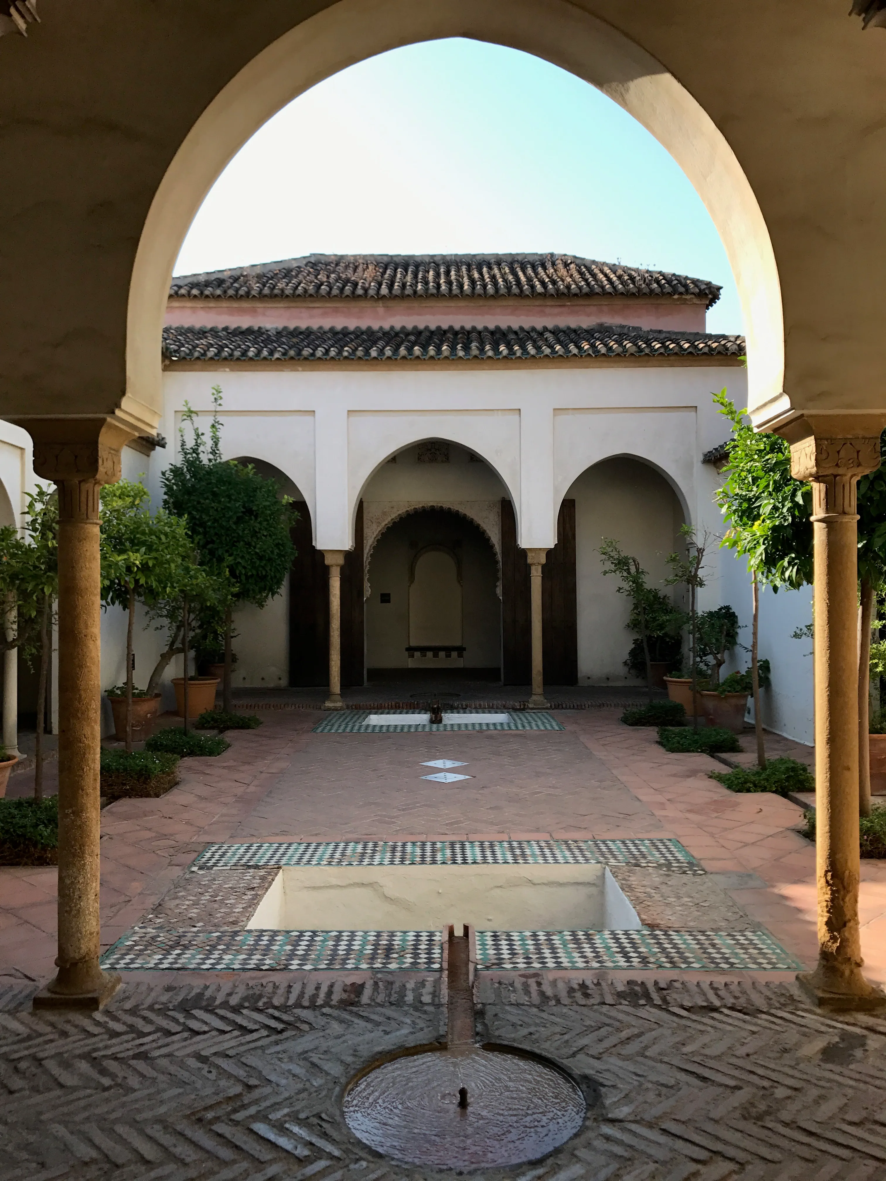 Patio soleado de la Alcazaba de Málaga visto a través de un gran arco, con una fuente de azulejos, columnas y paredes blancas con aberturas arqueadas.