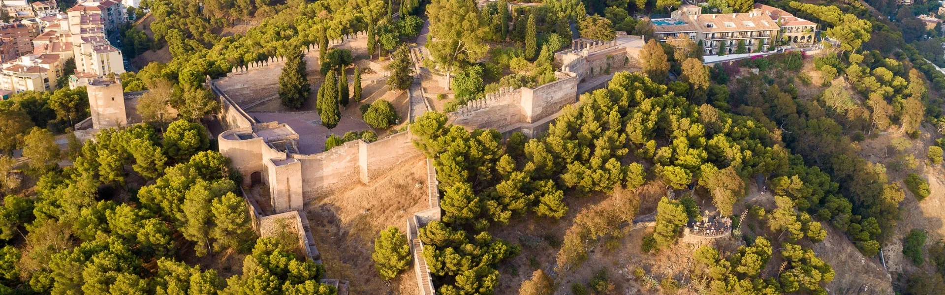 Vista panorámica aérea de las murallas y torres de la fortaleza de la Alcazaba, rodeada de árboles verdes en una ladera de Málaga.