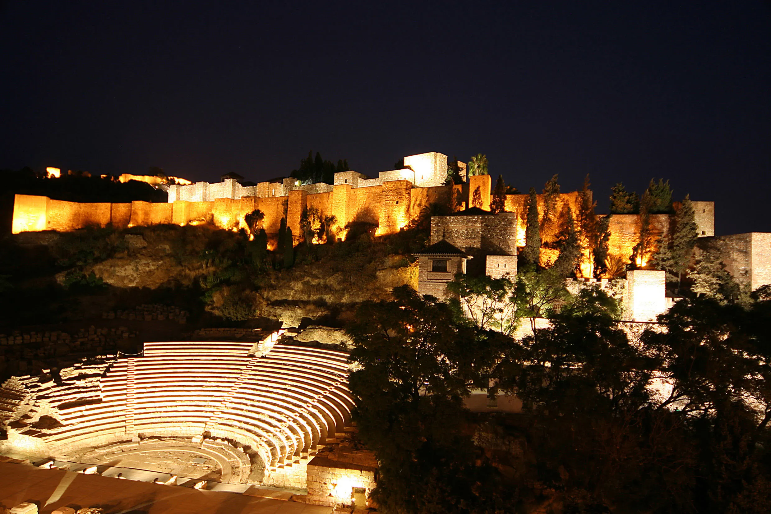 Vista nocturna de las murallas iluminadas de la fortaleza de la Alcazaba sobre una colina, por encima del iluminado Teatro Romano de Málaga.