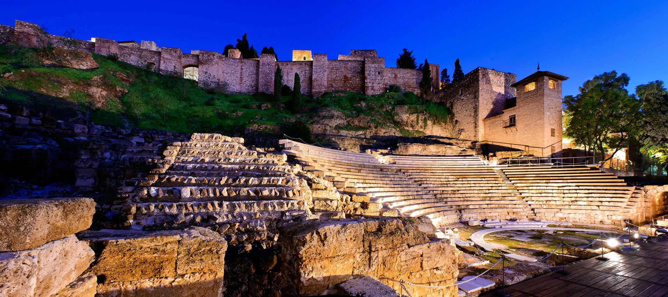 El Teatro Romano de Málaga iluminado al anochecer, con la alcazaba alzándose sobre la colina bajo un cielo crepuscular de color azul intenso.