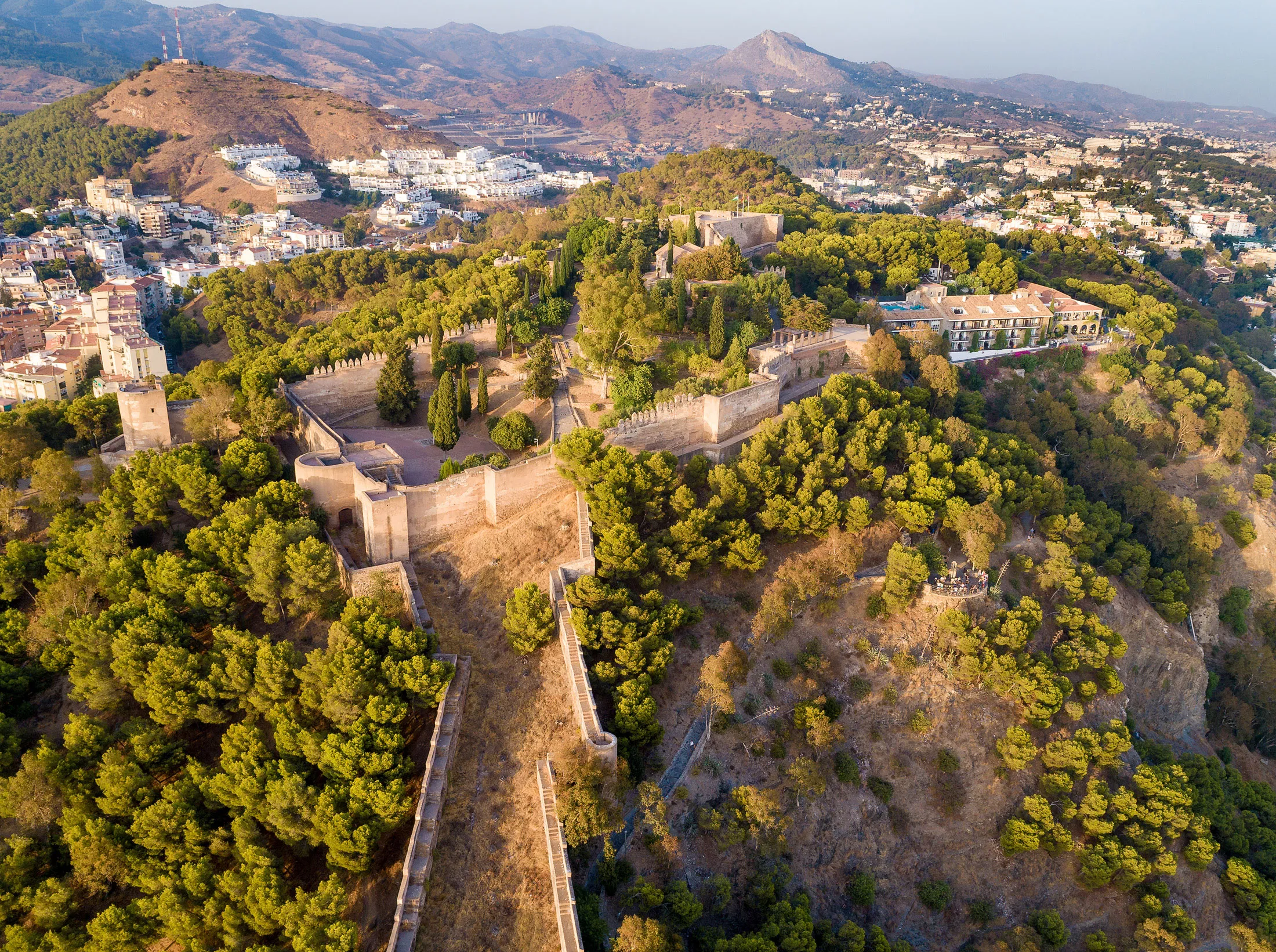 Vista aérea de las murallas de la fortaleza de la Alcazaba serpenteando por una ladera cubierta de pinos sobre Málaga, con la ciudad y las montañas visibles en el fondo.