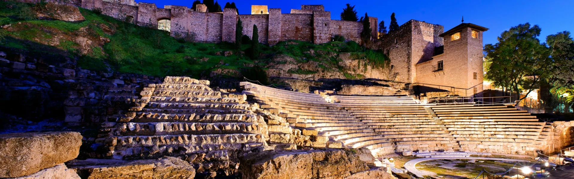 La fortaleza iluminada de la Alcazaba se alza al anochecer sobre el antiguo Teatro Romano de Málaga, con las gradas de piedra en primer plano y las murallas defensivas brillando contra el cielo azul profundo.