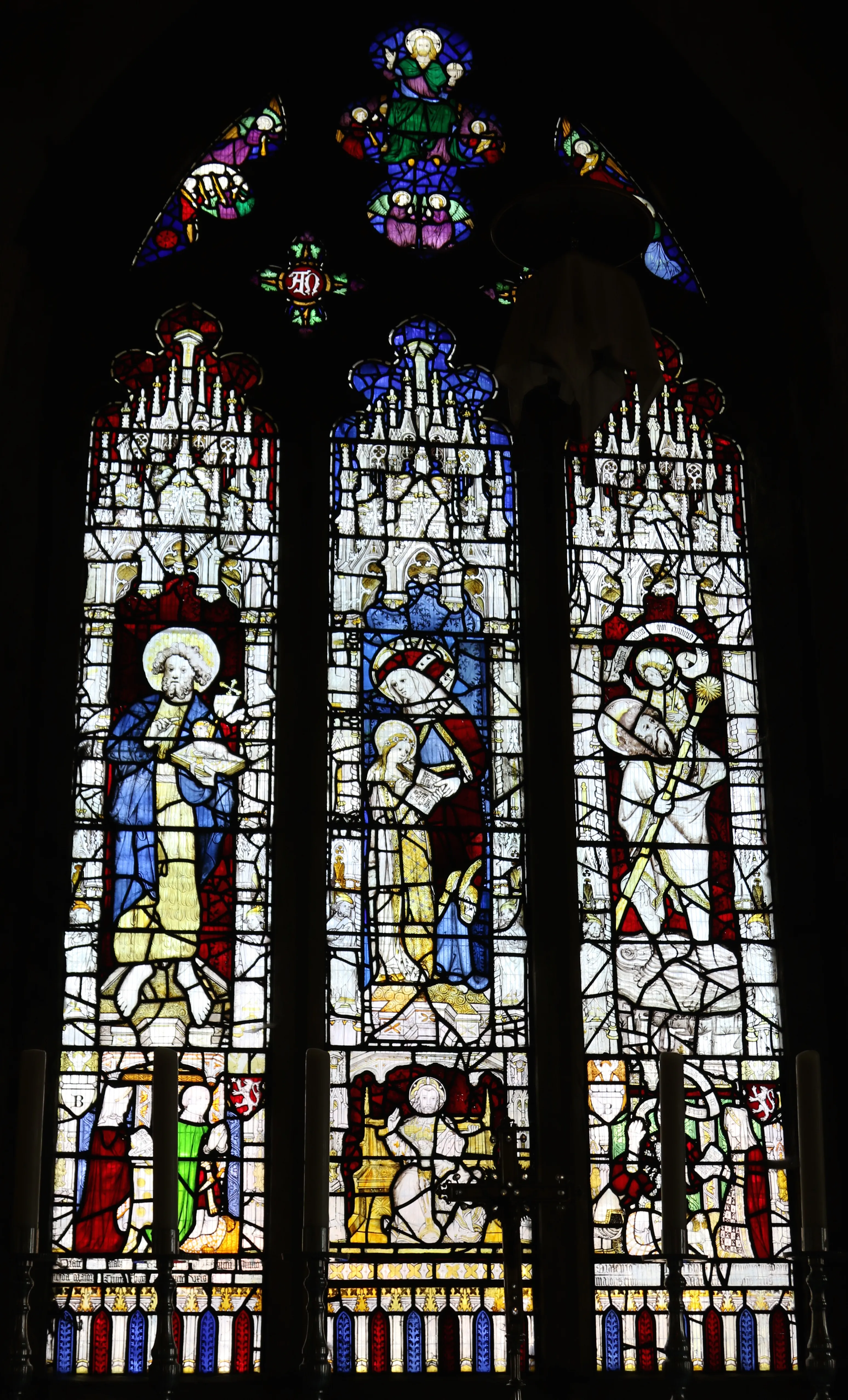 Three-panel medieval stained glass window depicting religious figures in blue, red, and yellow glass with decorative tracery at the top