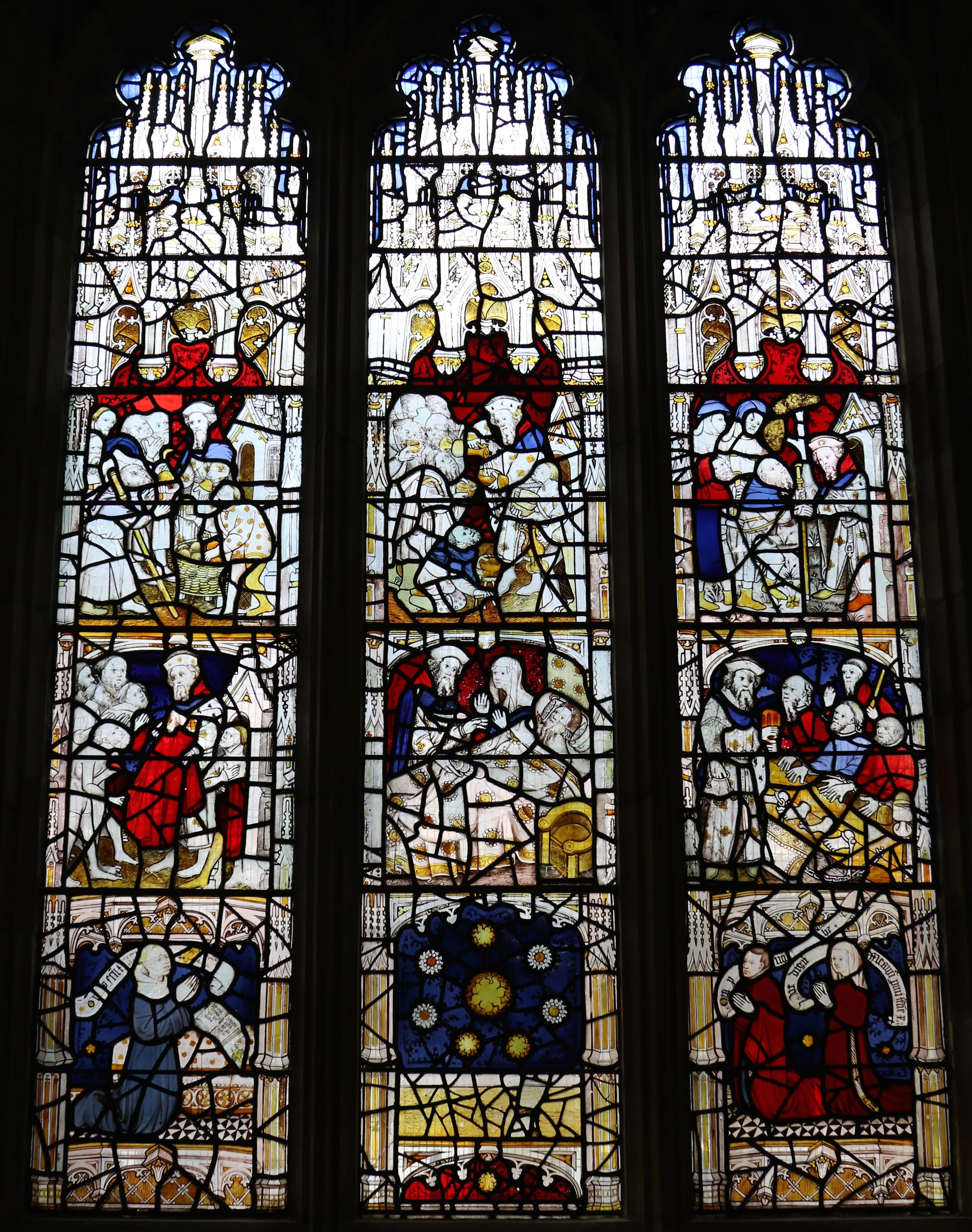 Three-panel medieval stained glass window depicting the Corporal Acts of Mercy with figures in red, blue, and white robes arranged in narrative scenes.