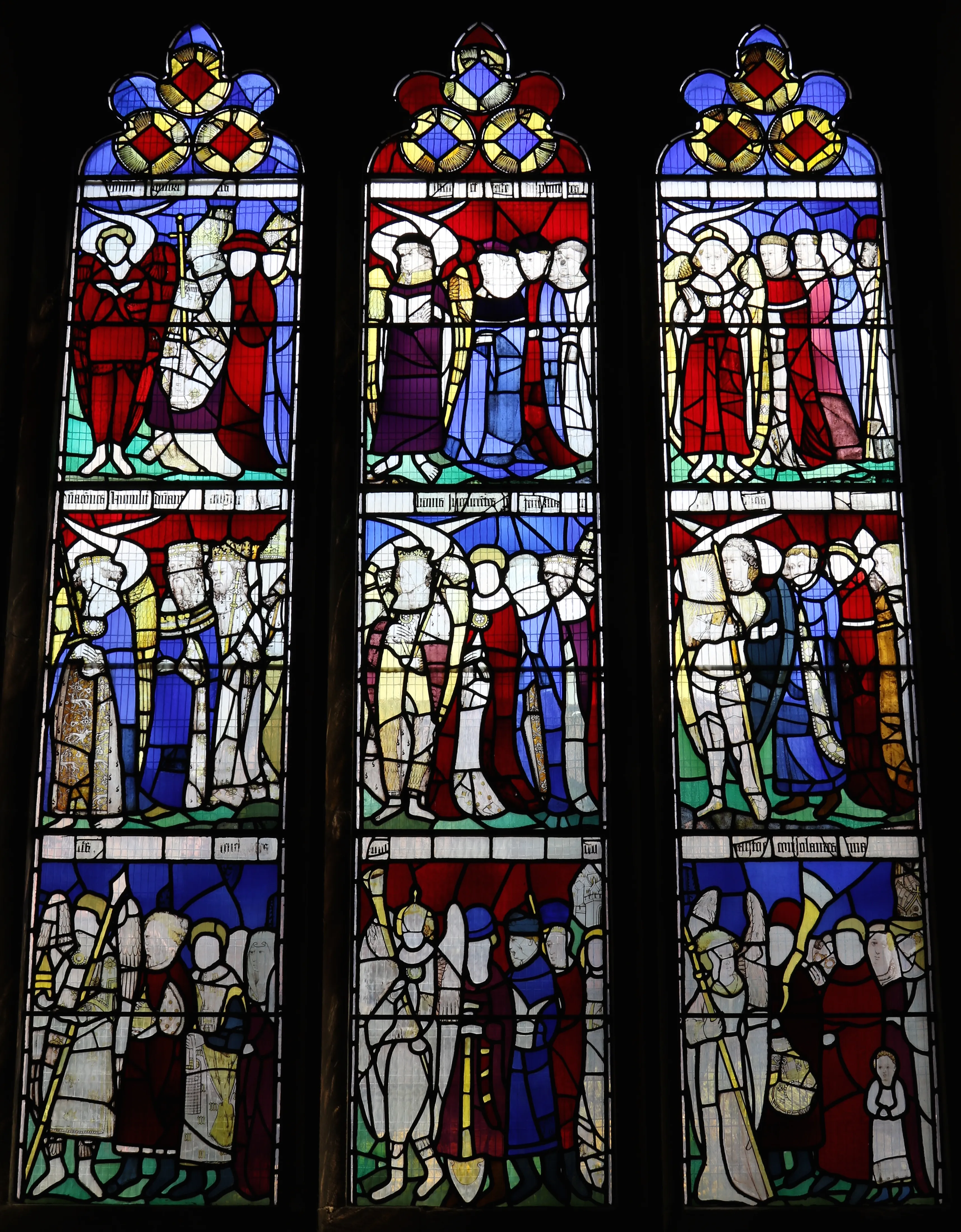 Three-panel medieval stained glass window depicting nine orders of angels in colorful robes of red, blue, purple, and yellow, with Gothic arched tops and Latin inscriptions between panels