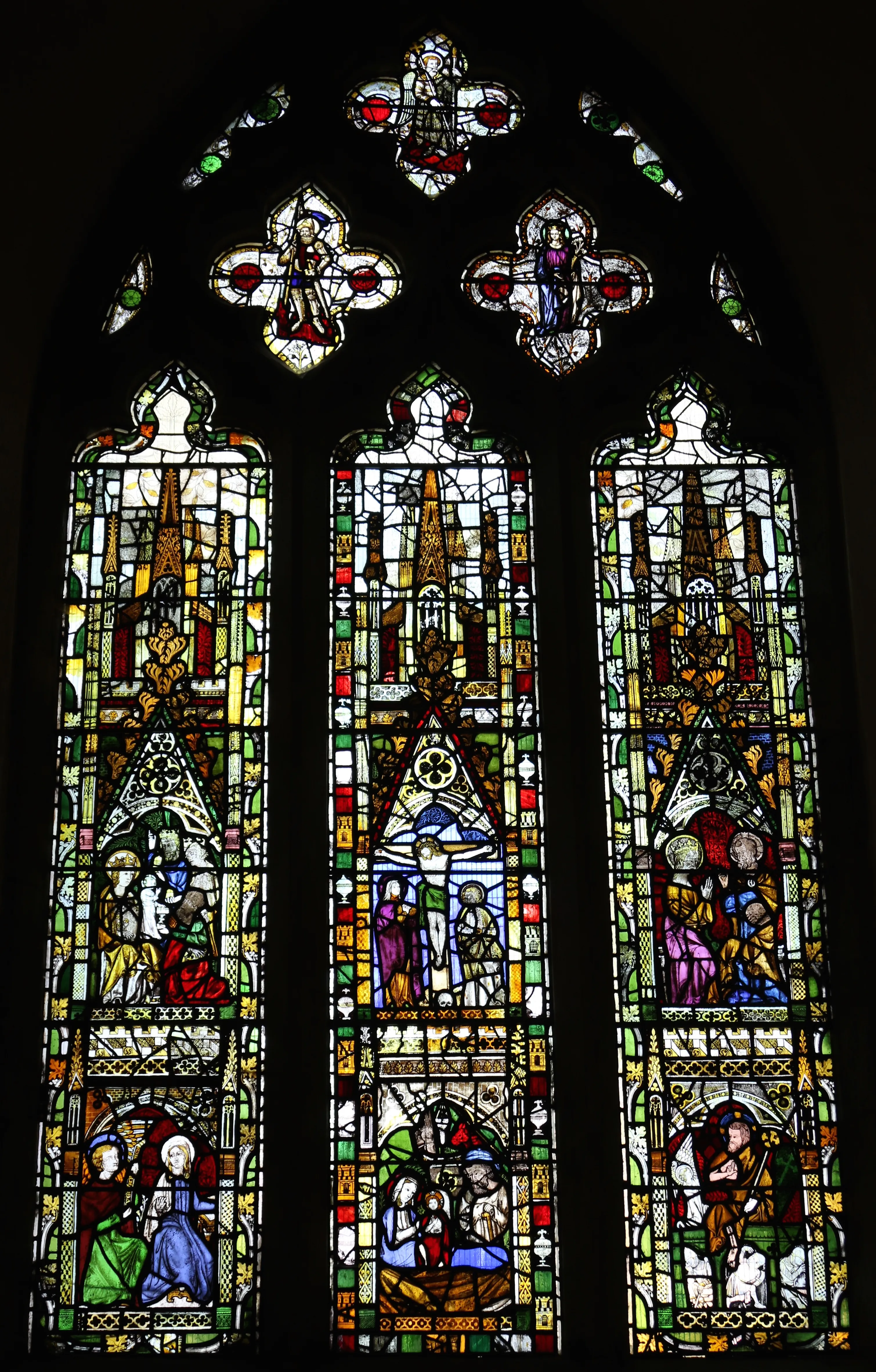 Three-panel medieval stained glass window with Gothic tracery, depicting religious scenes in vibrant red, blue, green, and yellow glass with figures in arched compartments