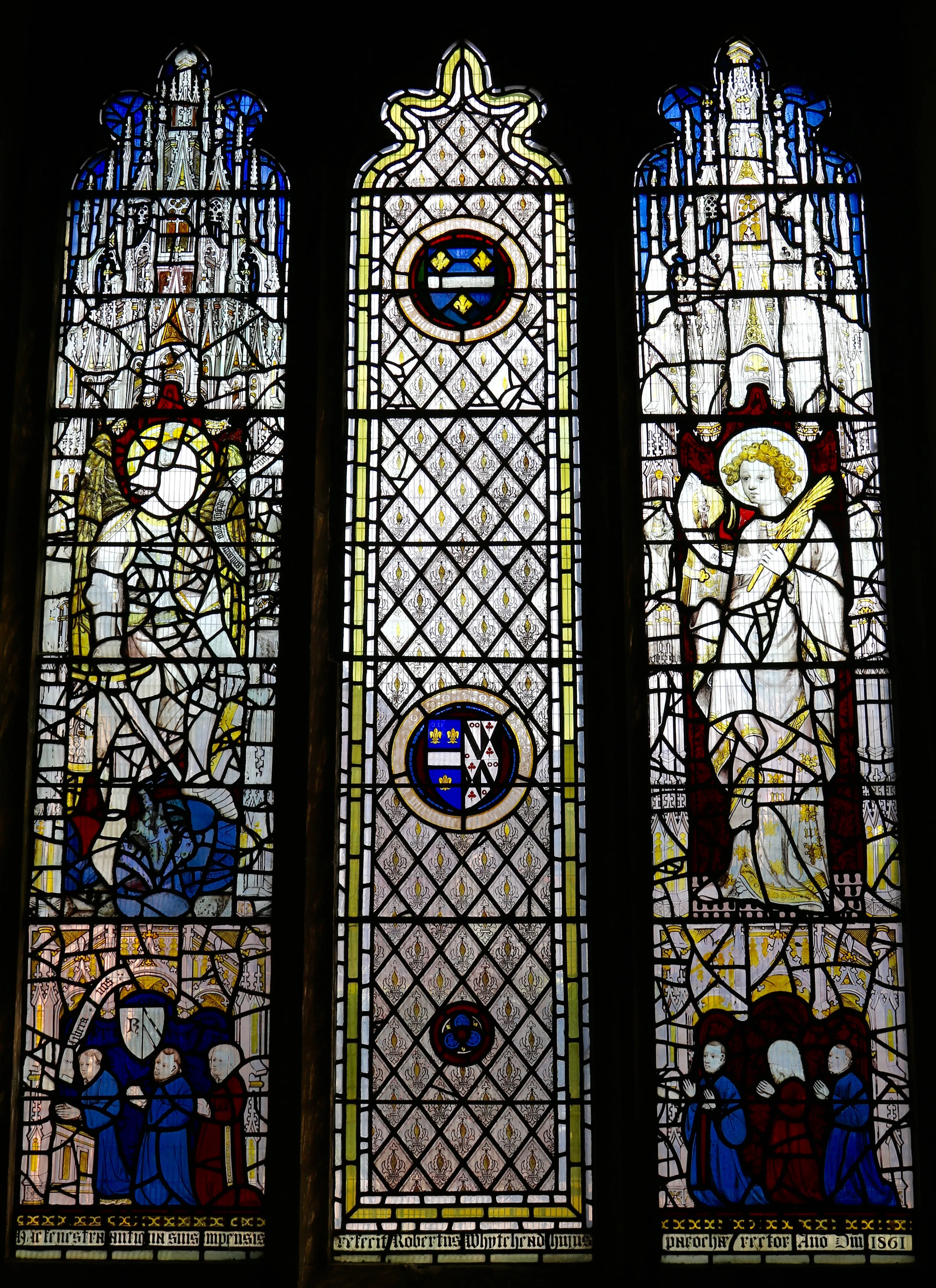 Three-panel medieval stained glass window showing haloed religious figures in the left and right panels, heraldic shields in roundels in the central panel, and small donor figures at the bottom of each side panel
