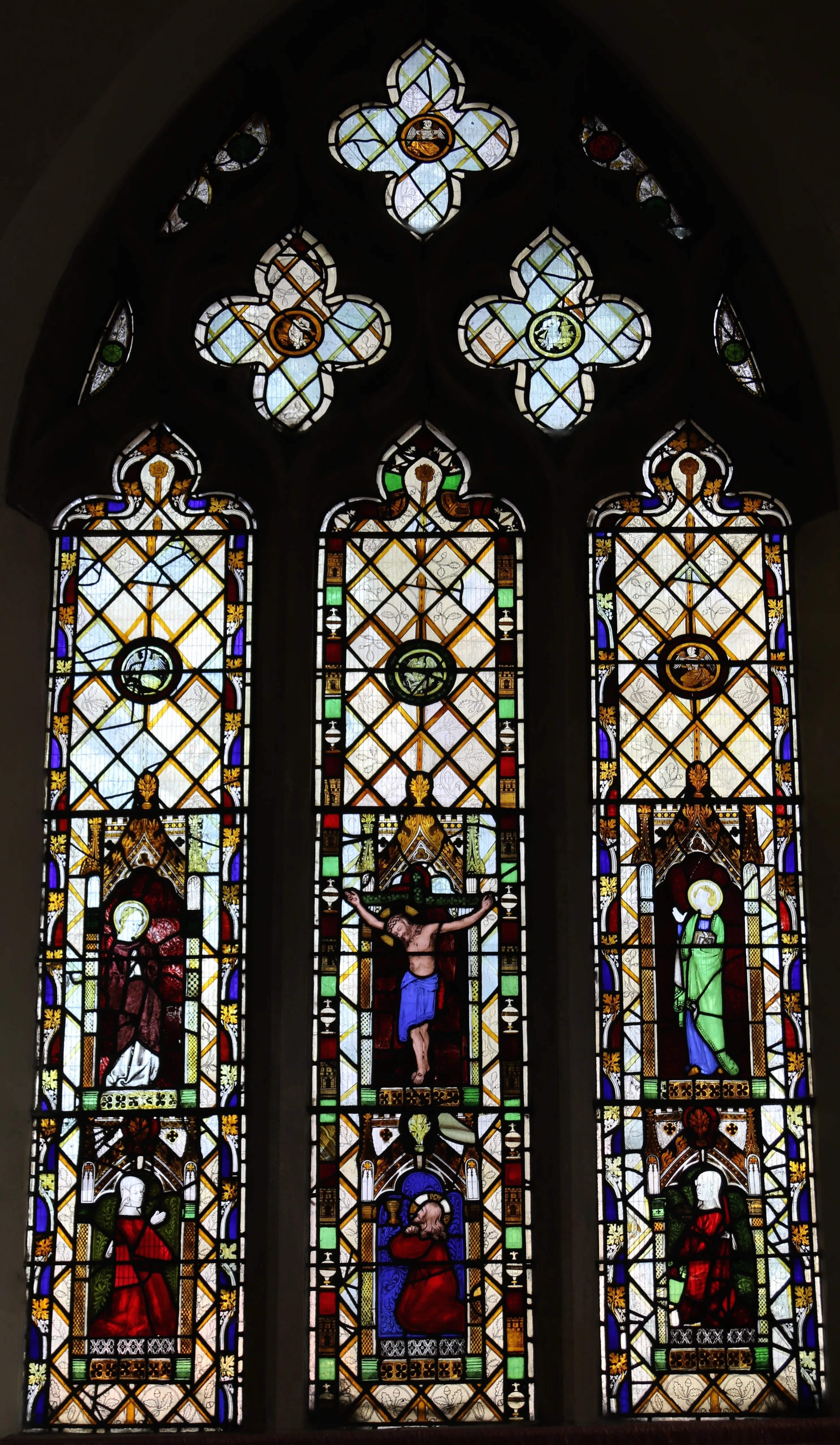 Tall three-paneled stained glass window with a pointed arch top featuring quatrefoil tracery, depicting a central crucifixion scene flanked by standing religious figures in red and green robes.