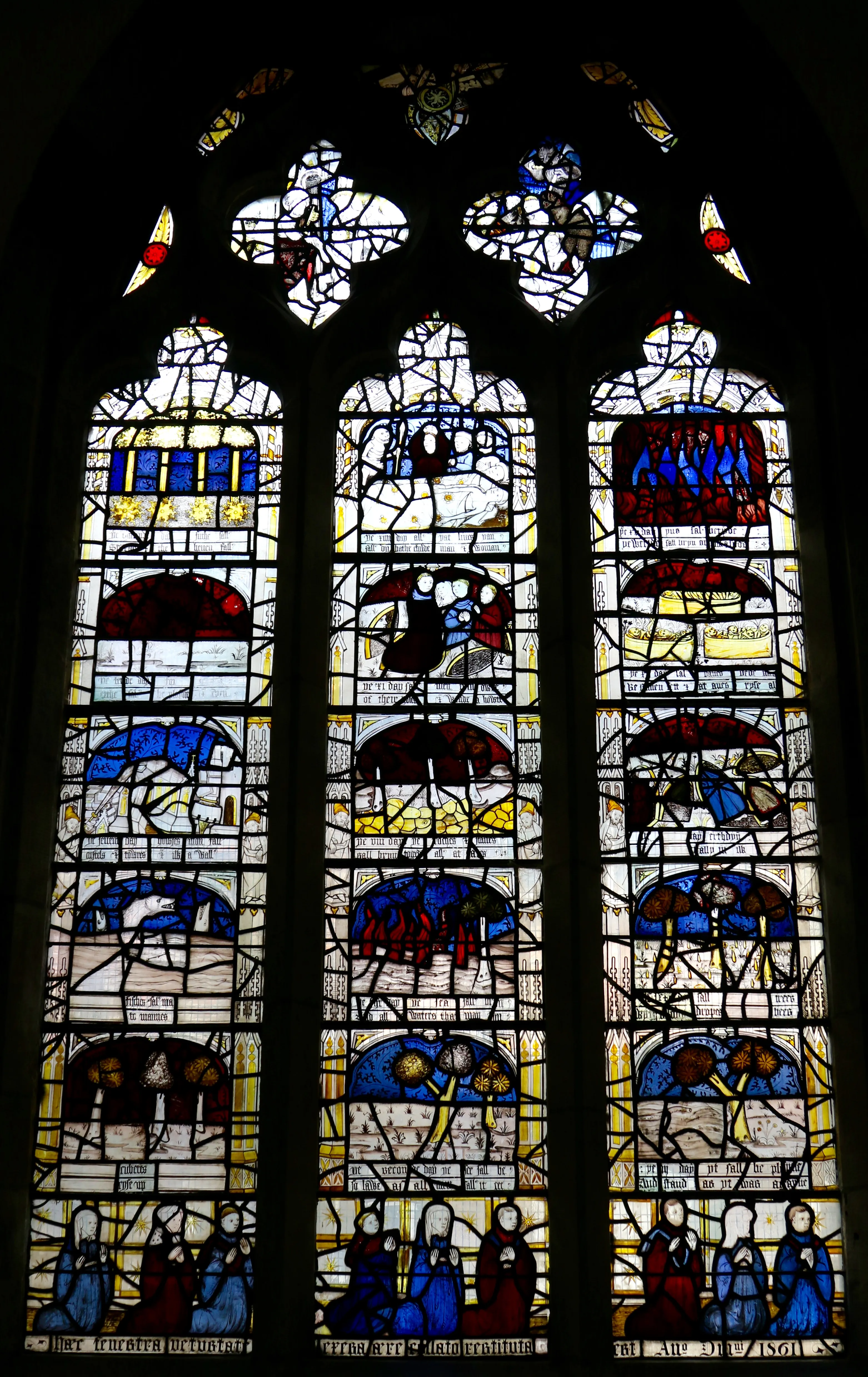 Three-panel Gothic stained glass window with multiple narrative scenes in blue, red, white, and yellow, featuring figures at the bottom of each panel