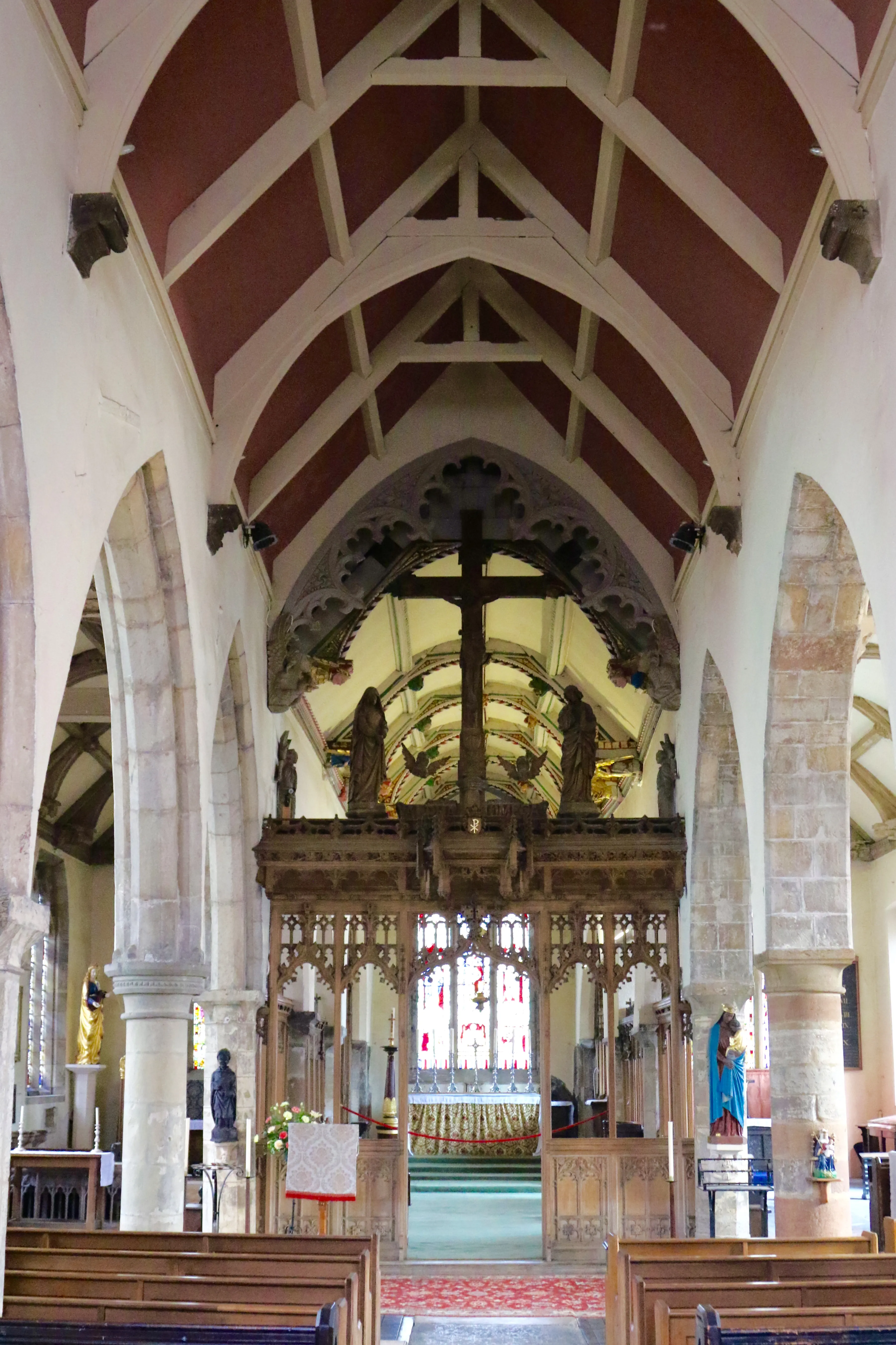 Interior view of All Saints Church showing the nave, chancel, and a wooden rood screen with a crucifix and statues beneath a red and white timber ceiling.