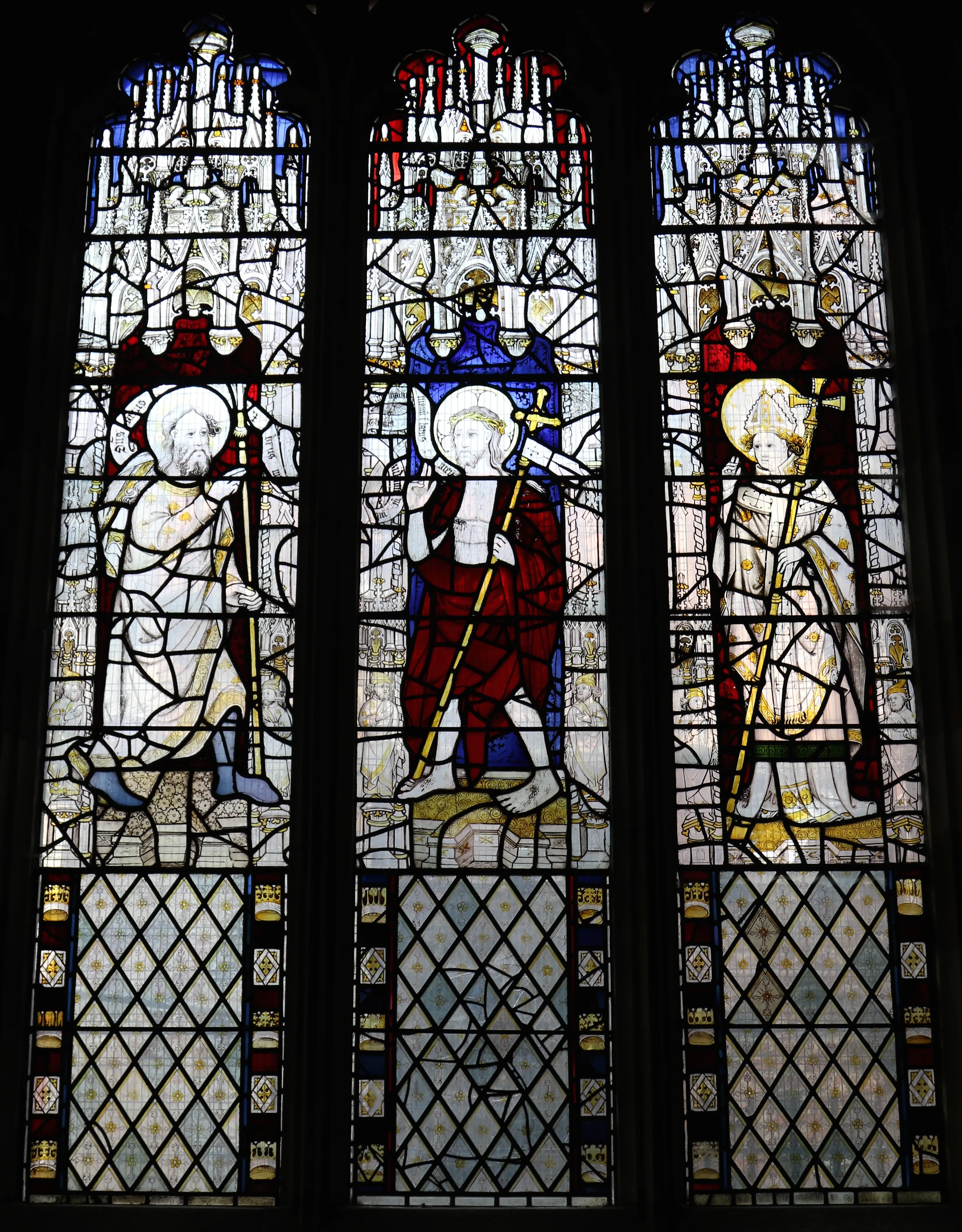 Three-panel Gothic stained glass window showing three haloed figures holding staffs, dressed in red, blue, and white robes, with diamond lattice patterns below.