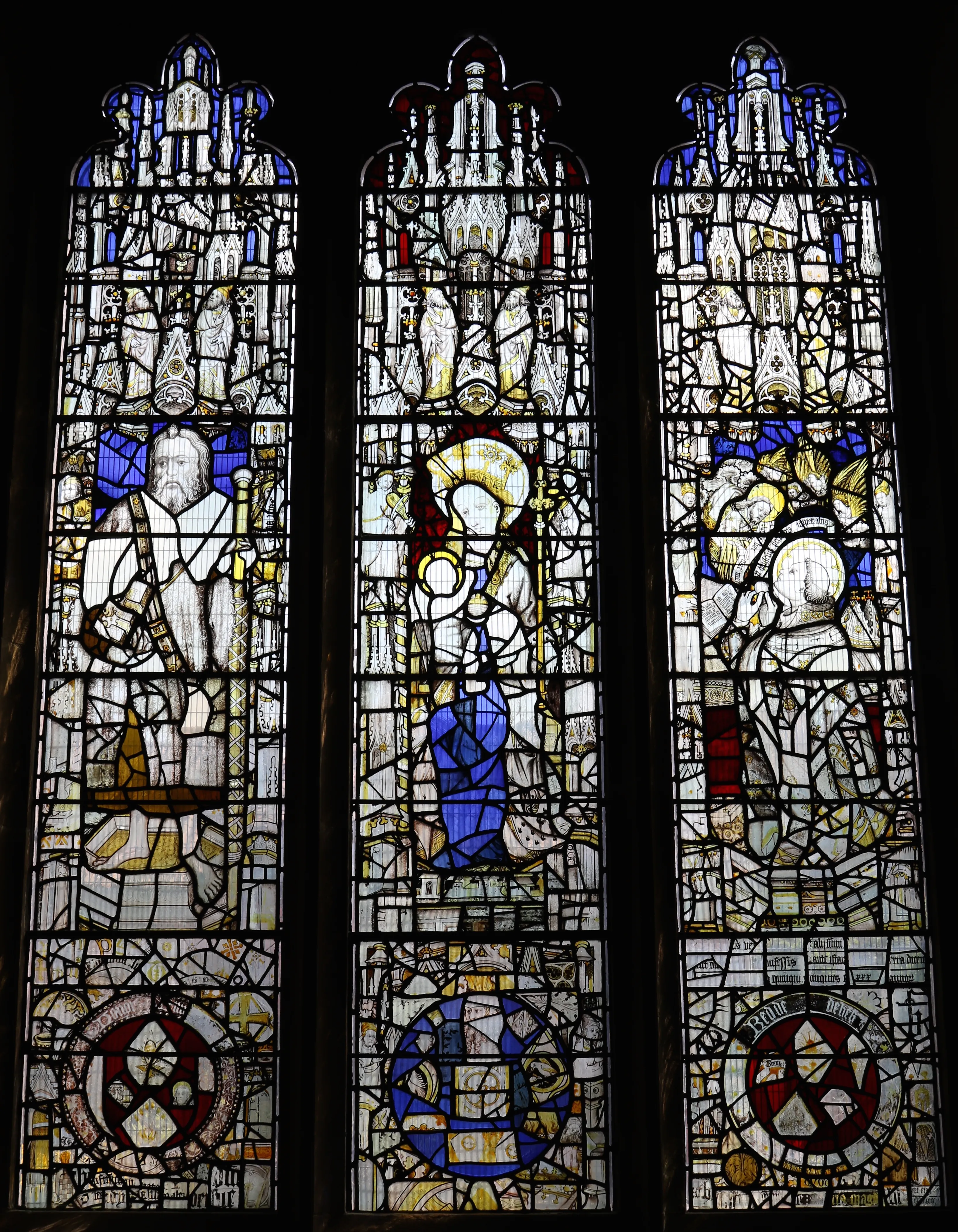 Three-panel stained glass window depicting robed religious figures with halos, topped with architectural canopies and featuring heraldic shields at the bottom