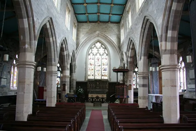 Interior de la nave de la All Saints Church que muestra arcos apuntados de piedra, bancos de madera a los lados de un pasillo con alfombra roja, un púlpito de madera y una vidriera al fondo bajo un techo pintado de azul