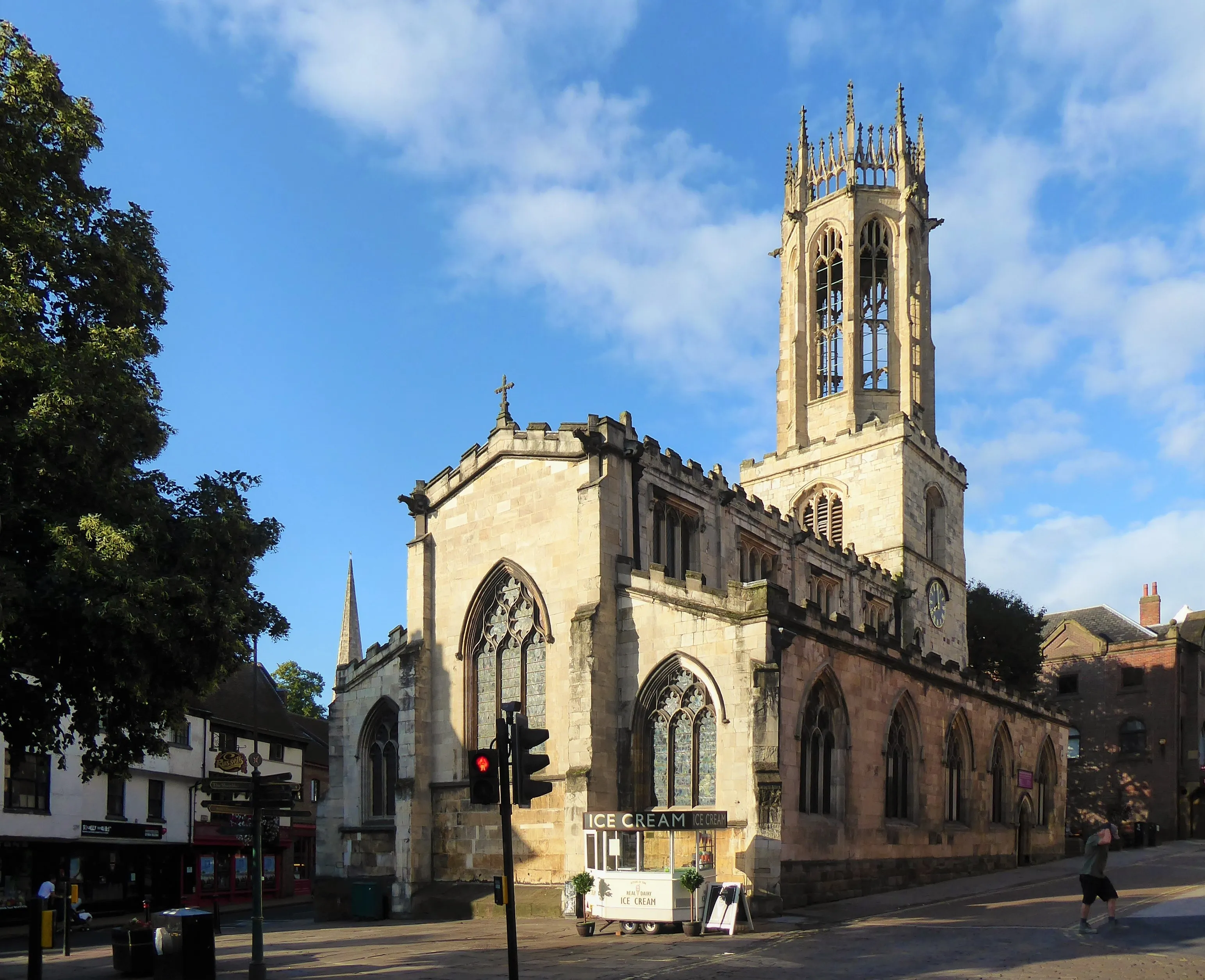 Vista exterior de la iglesia All Saints Church on Pavement en York, que destaca por su distintiva torre linterna medieval, su arquitectura gótica de piedra y sus ventanas arqueadas bajo un cielo azul.