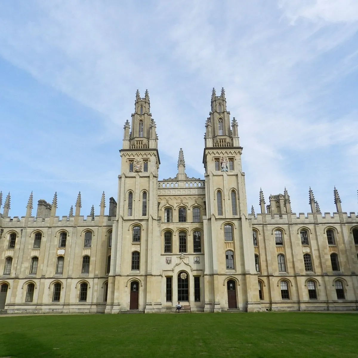 Gotycka fasada z dwiema wieżami All Souls College w Oksfordzie z ozdobnymi detalami kamieniarskimi, pinaklami i centralnym wejściem, na tle błękitnego nieba z zielonym trawnikiem na pierwszym planie.