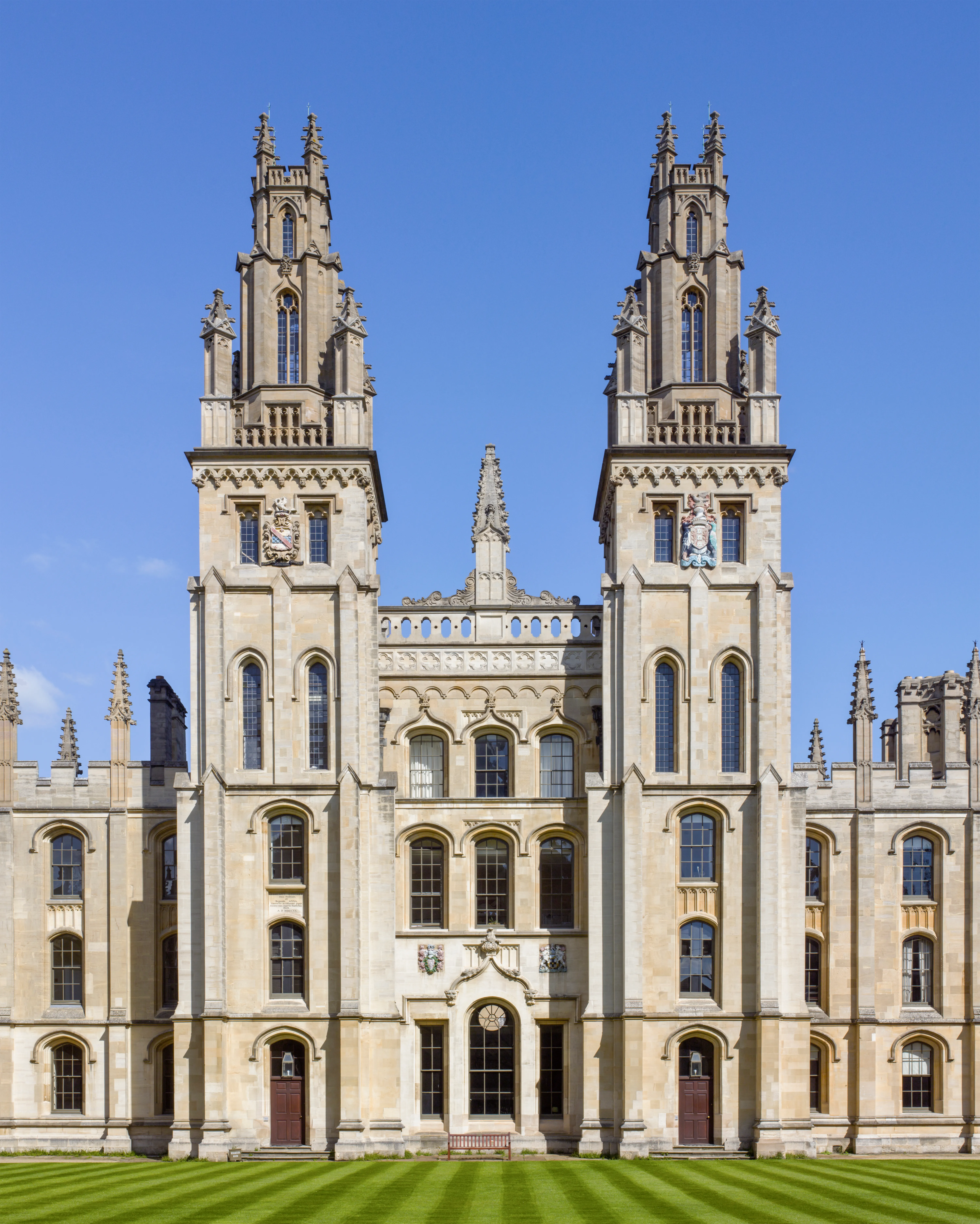 Bliźniacze gotyckie wieże All Souls College w Oksfordzie z ozdobną kamienną fasadą, łukowatymi oknami i pasiastym zielonym trawnikiem na pierwszym planie pod bezchmurnym niebem.