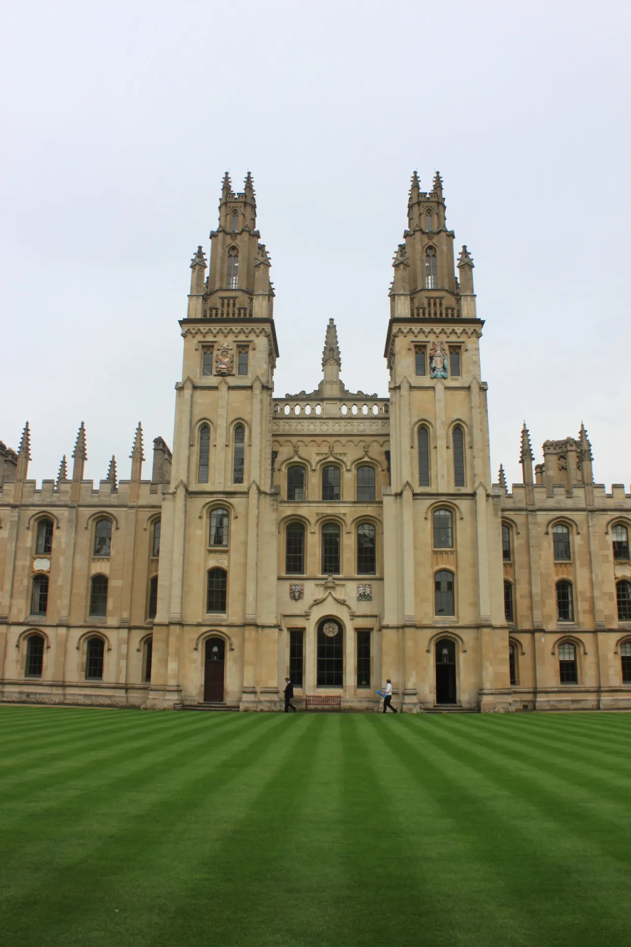 Gotycka kamienna fasada All Souls College z dwiema wieżami, łukowatymi oknami, sterczynami oraz pasiastym zielonym trawnikiem na pierwszym planie