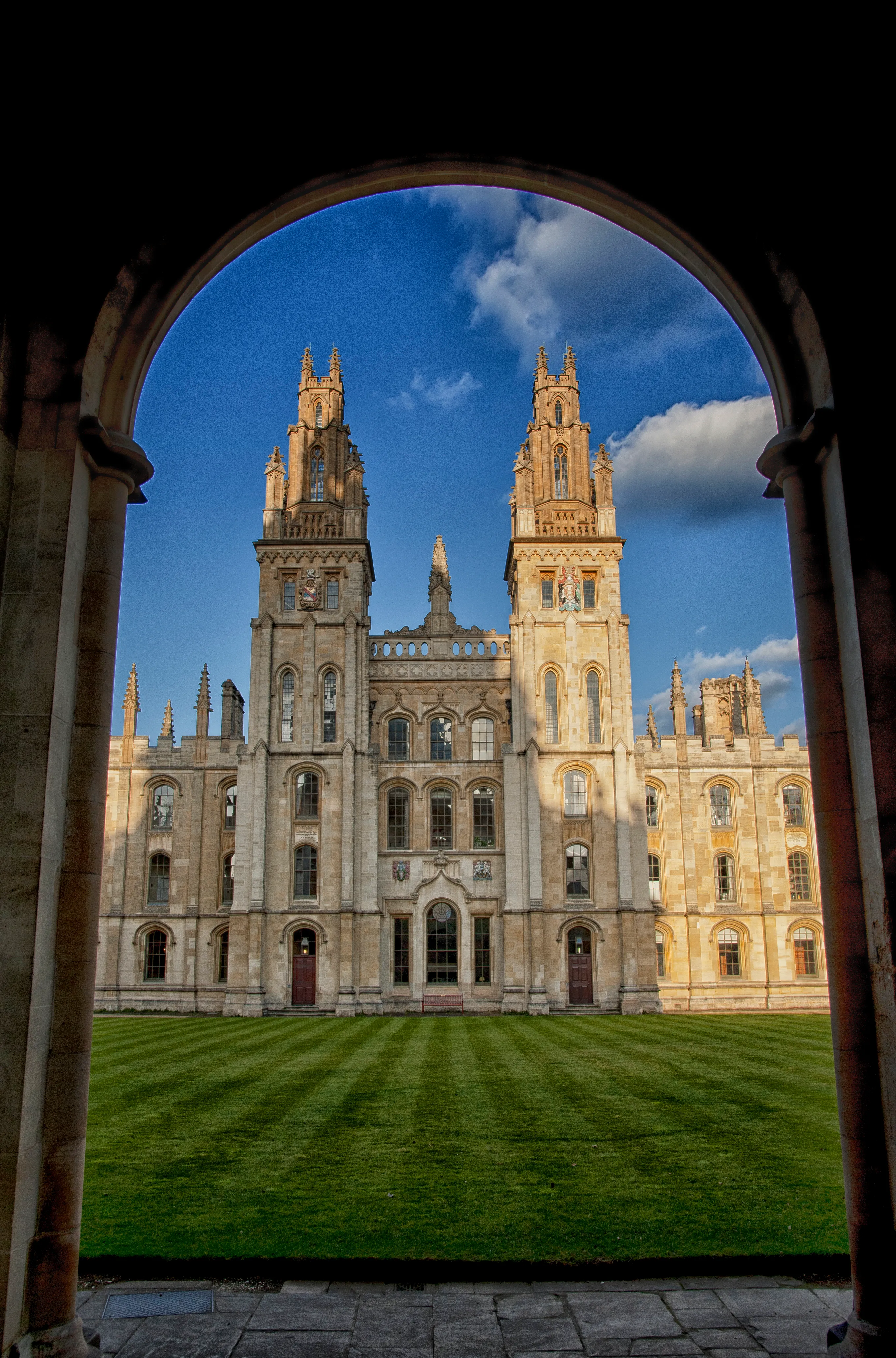Bliźniacze wieże All Souls College w Oksfordzie widziane przez kamienny łuk ponad pasiastym trawnikiem pod błękitnym niebem.