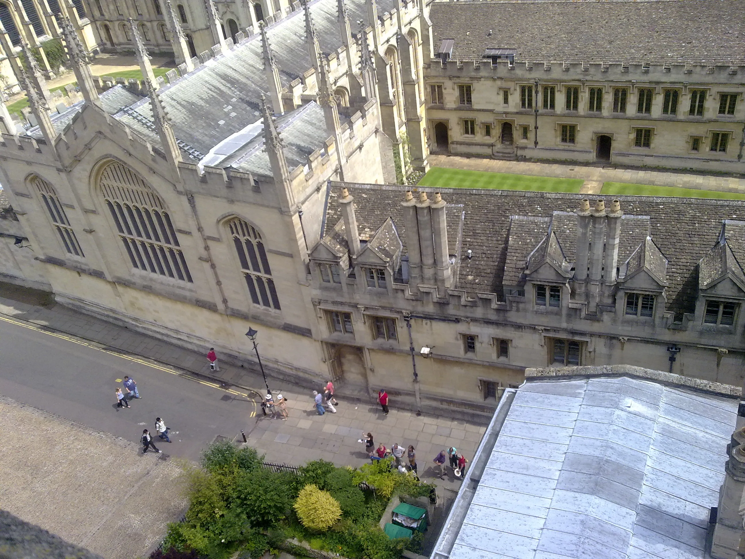 Widok z góry na kamienne dachy i gotycką architekturę All Souls College oraz Radcliffe Square, z pieszymi spacerującymi po chodniku poniżej.