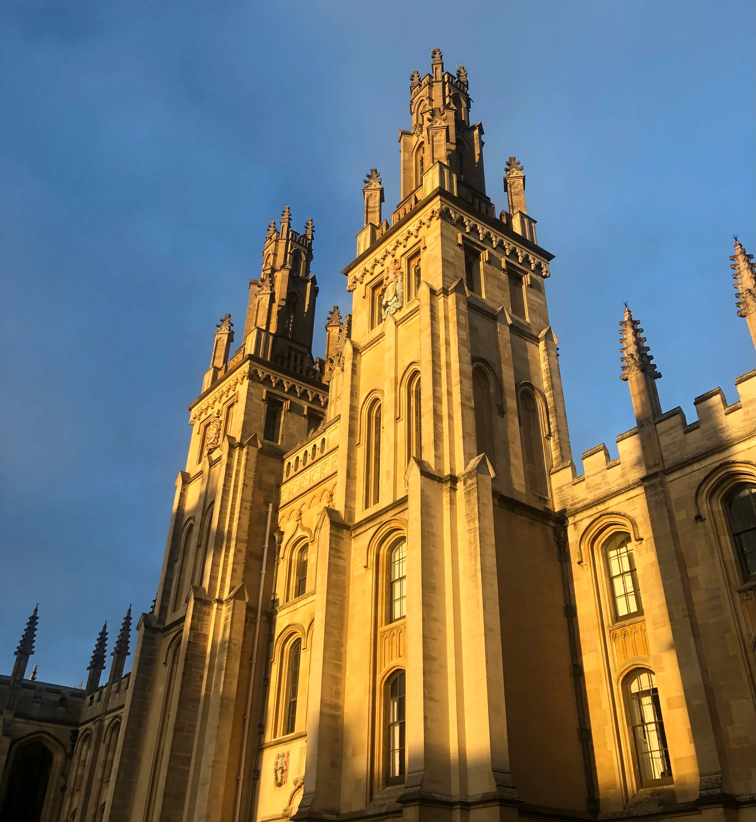 Bliźniacze gotyckie wieże All Souls College skąpane w ciepłym, złotym świetle słonecznym na tle bezchmurnego błękitnego nieba, z ozdobnymi pinaklami i kamienną fasadą
