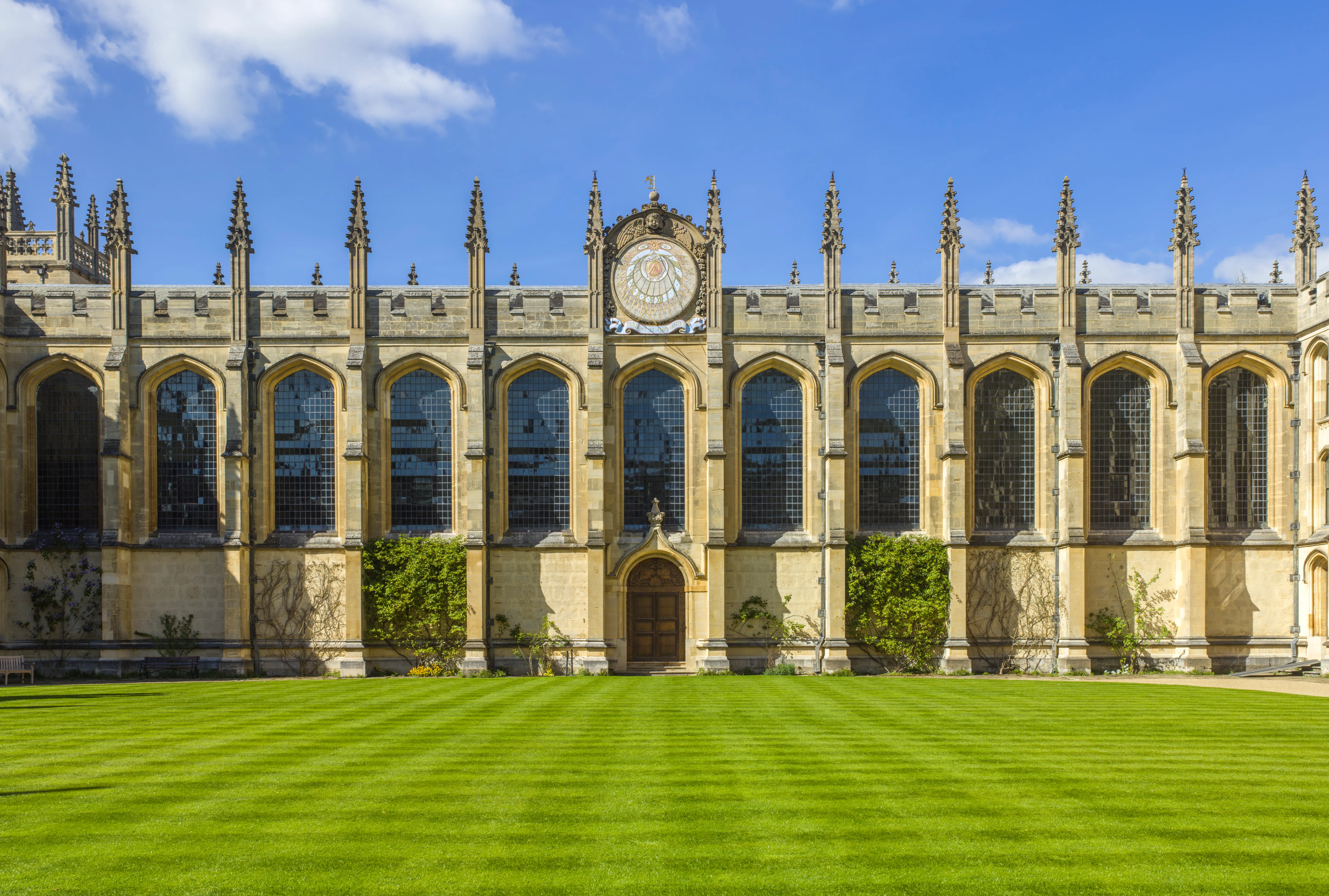 Gotycka kamienna fasada biblioteki Codrington Library w All Souls College z centralnym zegarem, wysokimi łukowymi oknami, pinaklami i zadbanym zielonym trawnikiem pod błękitnym niebem