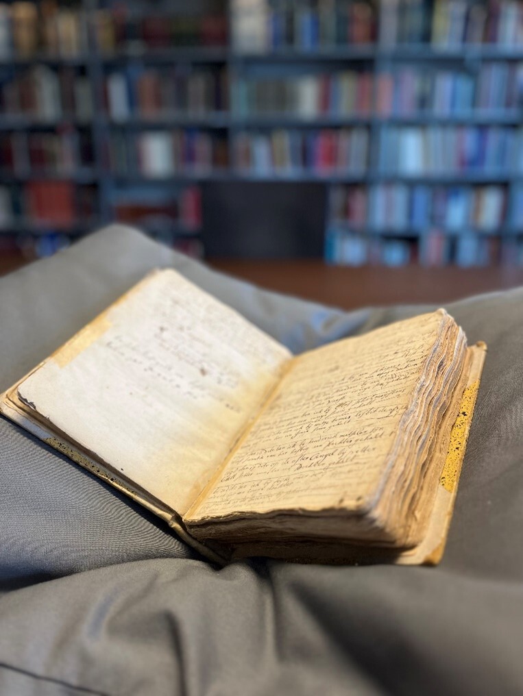 Open oude manuscript met handgeschreven tekst op vergeelde pagina's rustend op grijze stof, boekenplanken op de achtergrond